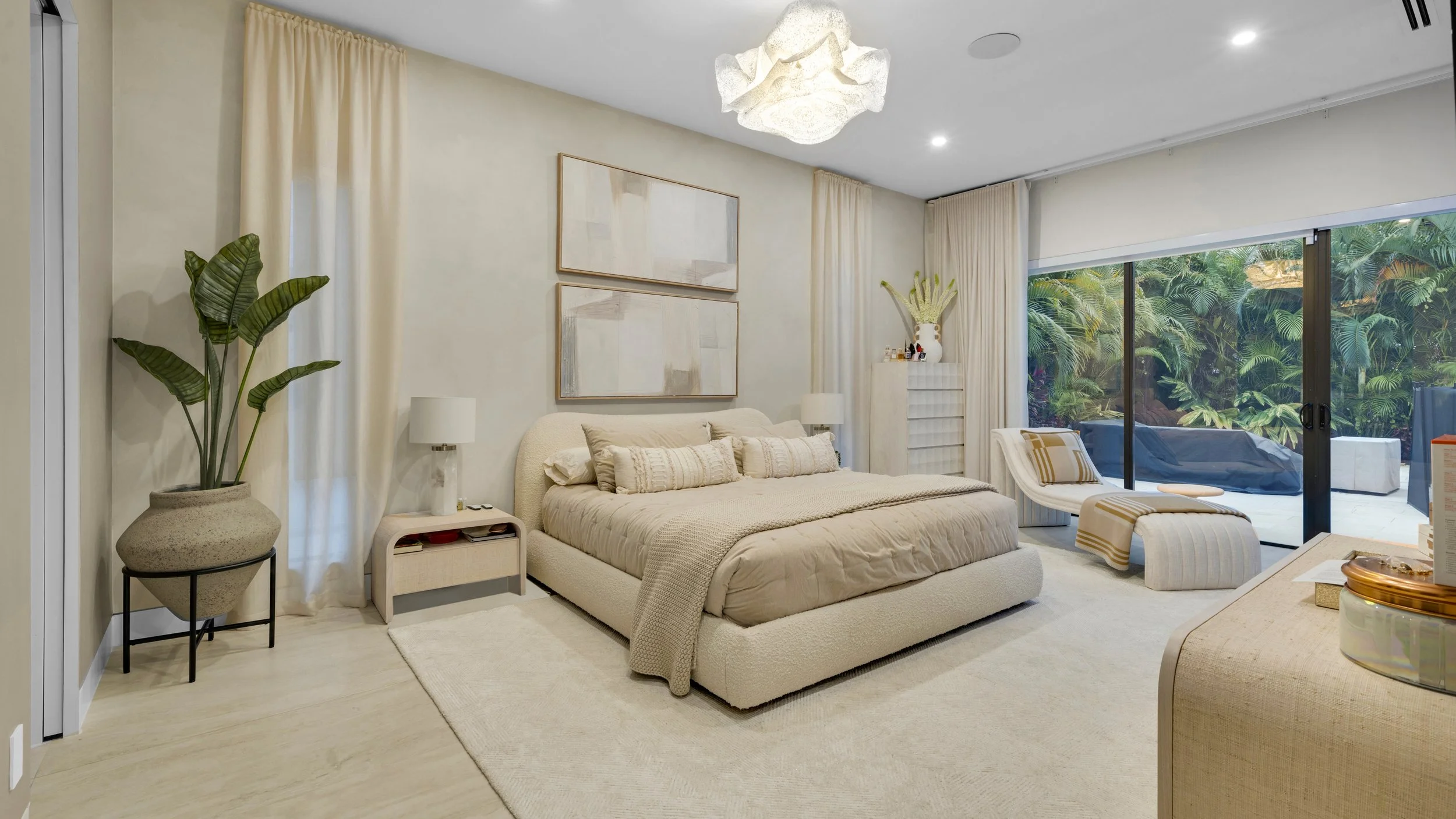 A contemporary bedroom with a large bed, beige bedding, and matching pillows, flanked by two small bedside tables with lamps. A tall potted plant is in the corner, next to curtain-covered window. Artwork hangs above the bed. There is a white lounge c