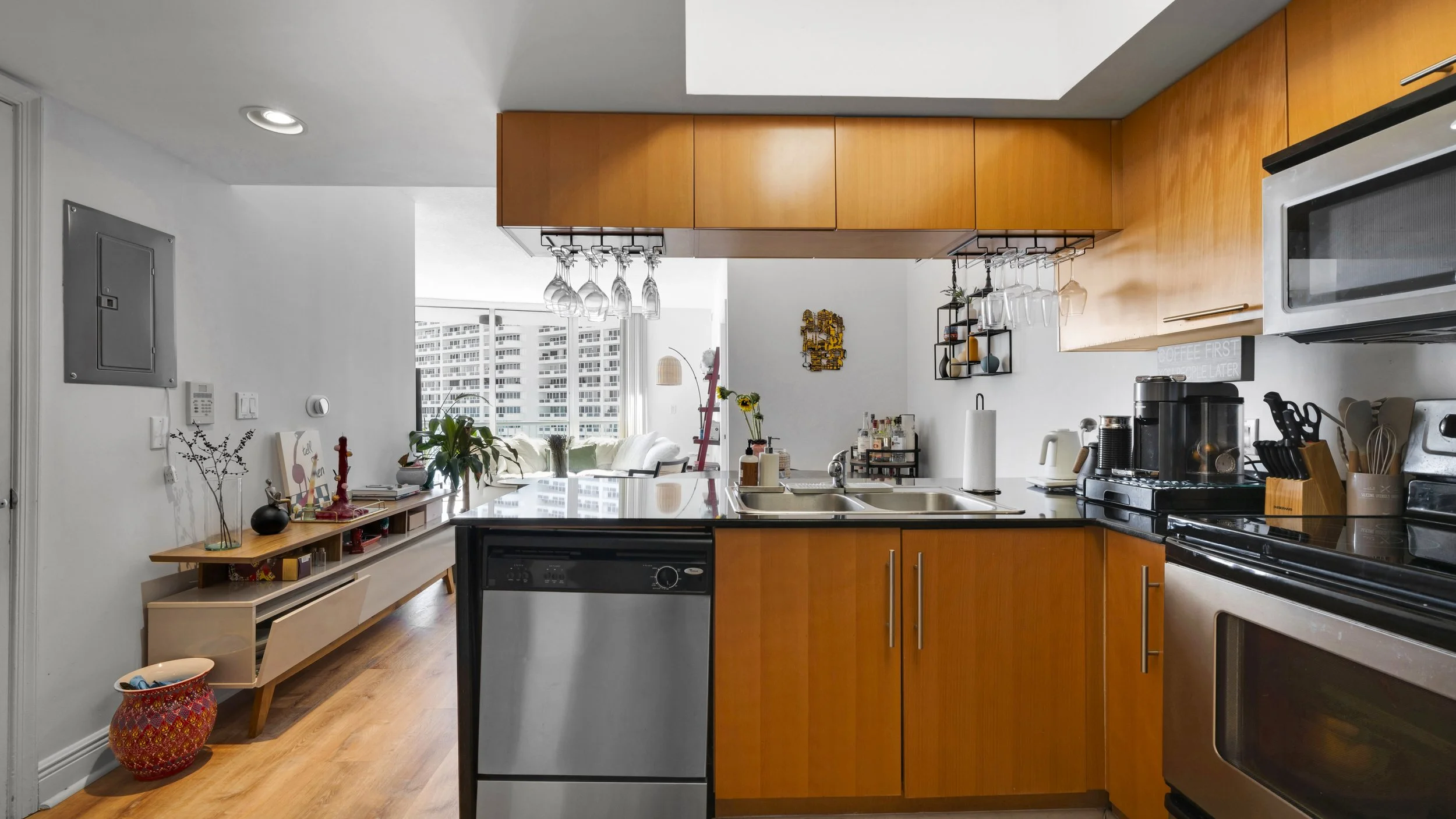 Modern kitchen with wooden cabinets, stainless steel appliances, and a view of a living room with large windows and city buildings.