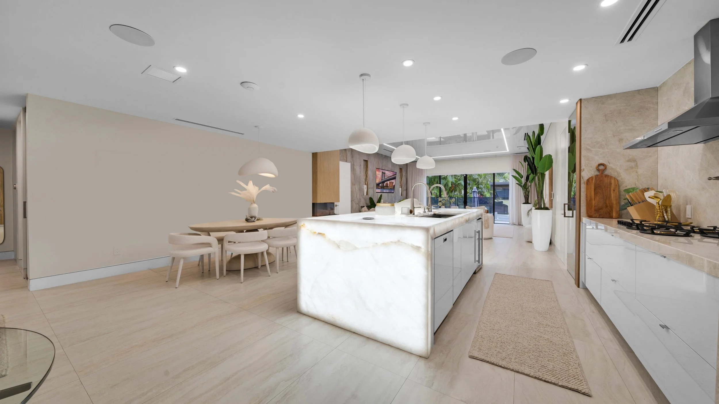 Modern open-concept kitchen with a marble island, white cabinets, and a dining area with a round table and six chairs. Large potted plants and sliding glass doors leading to an outdoor area.