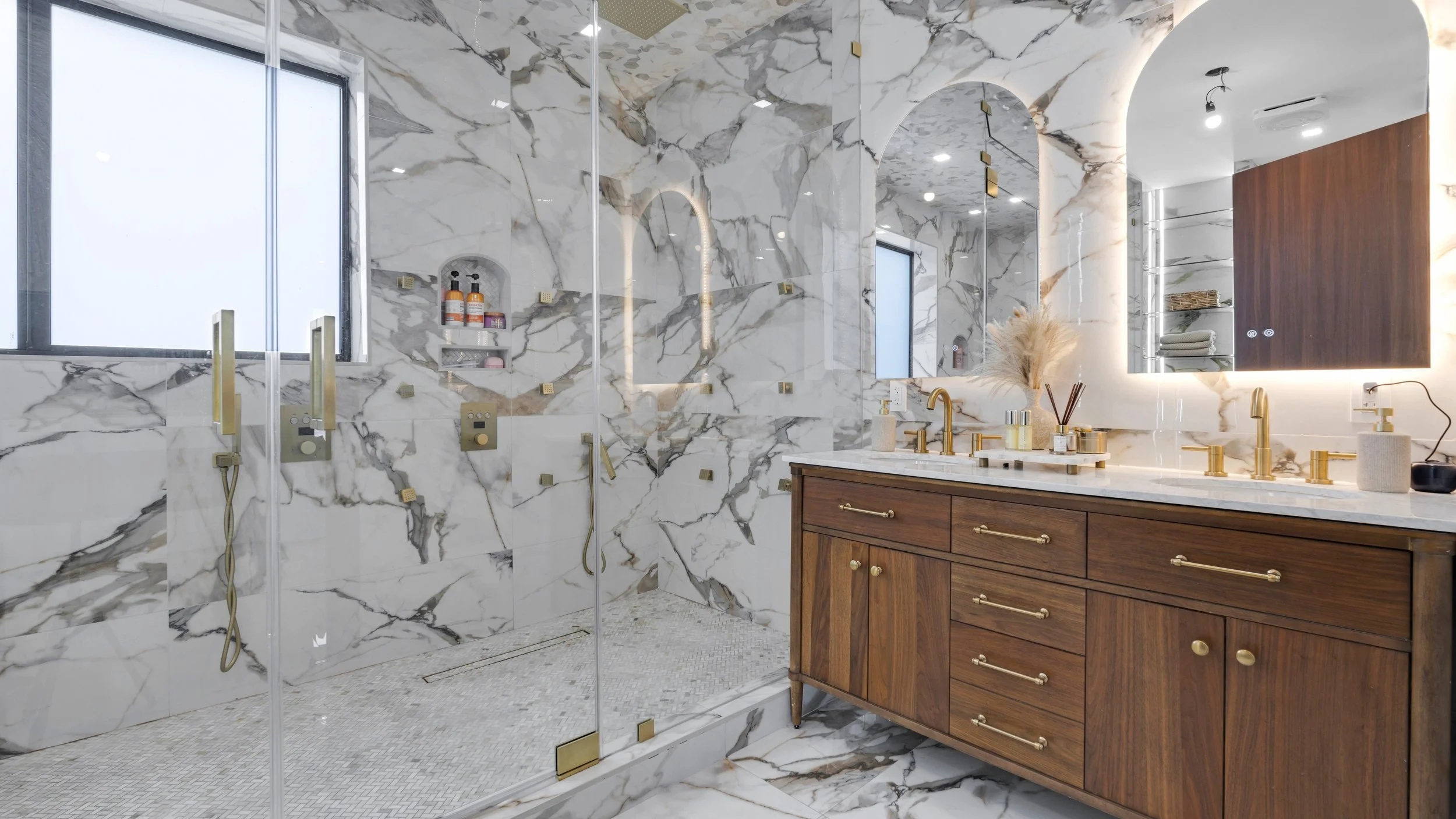 A luxurious bathroom with marble walls and floors, a wooden vanity with gold hardware, a large mirror with backlighting, and a walk-in shower with glass doors and gold fixtures.