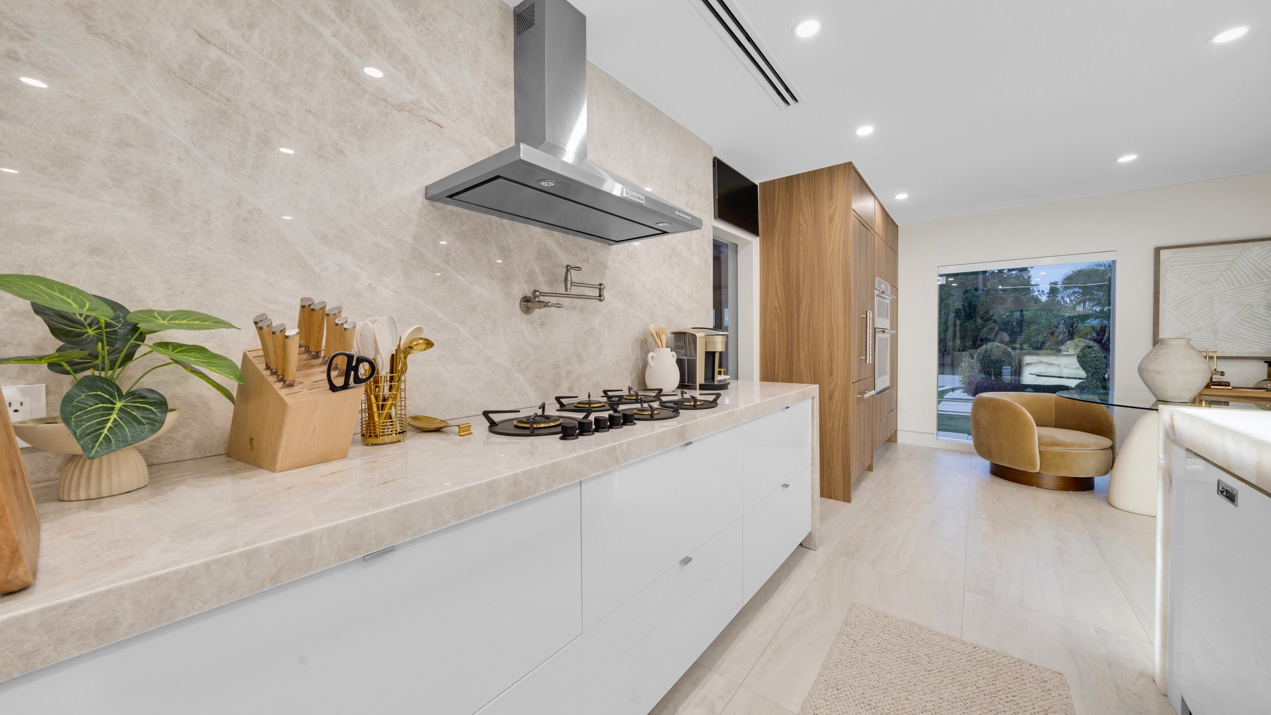 Modern kitchen with white cabinetry, beige marble countertops, a stainless steel range hood, and a built-in oven. There are plants, kitchen utensils, and a coffee maker on the countertop. A cozy seating area with a tan armchair and decorative vases i