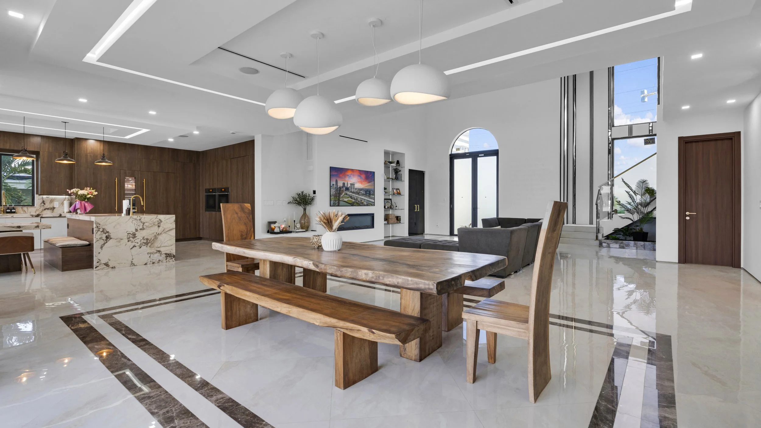 Modern open-concept living and dining area with wooden furniture, white walls, and large windows, featuring a kitchen in the background.