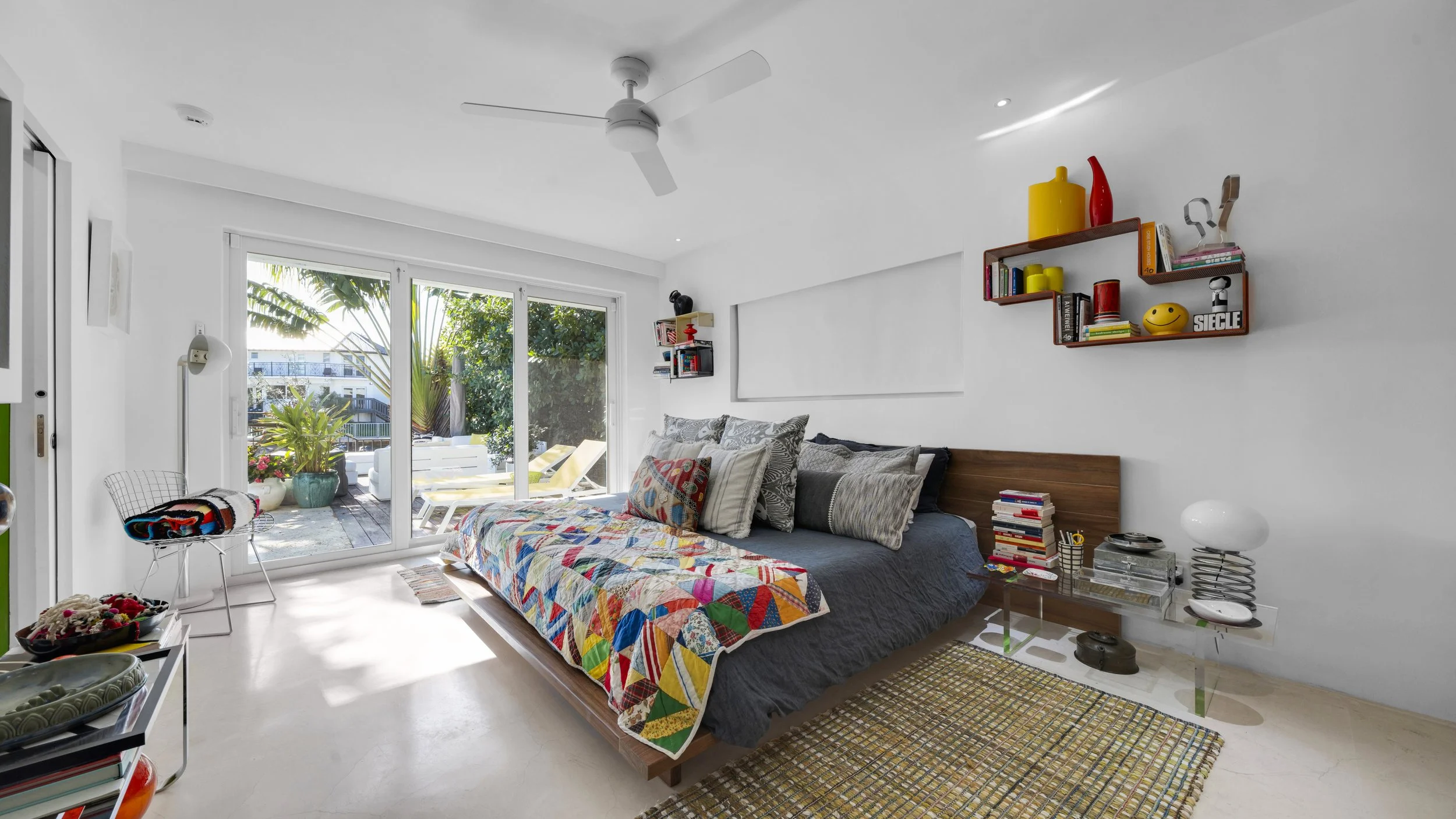 Bright bedroom with a large sliding glass door leading to a balcony with outdoor furniture and greenery. The bed has a colorful quilt and multiple pillows, with a wooden headboard and a transparent side table holding books and decor items. Wall-mount