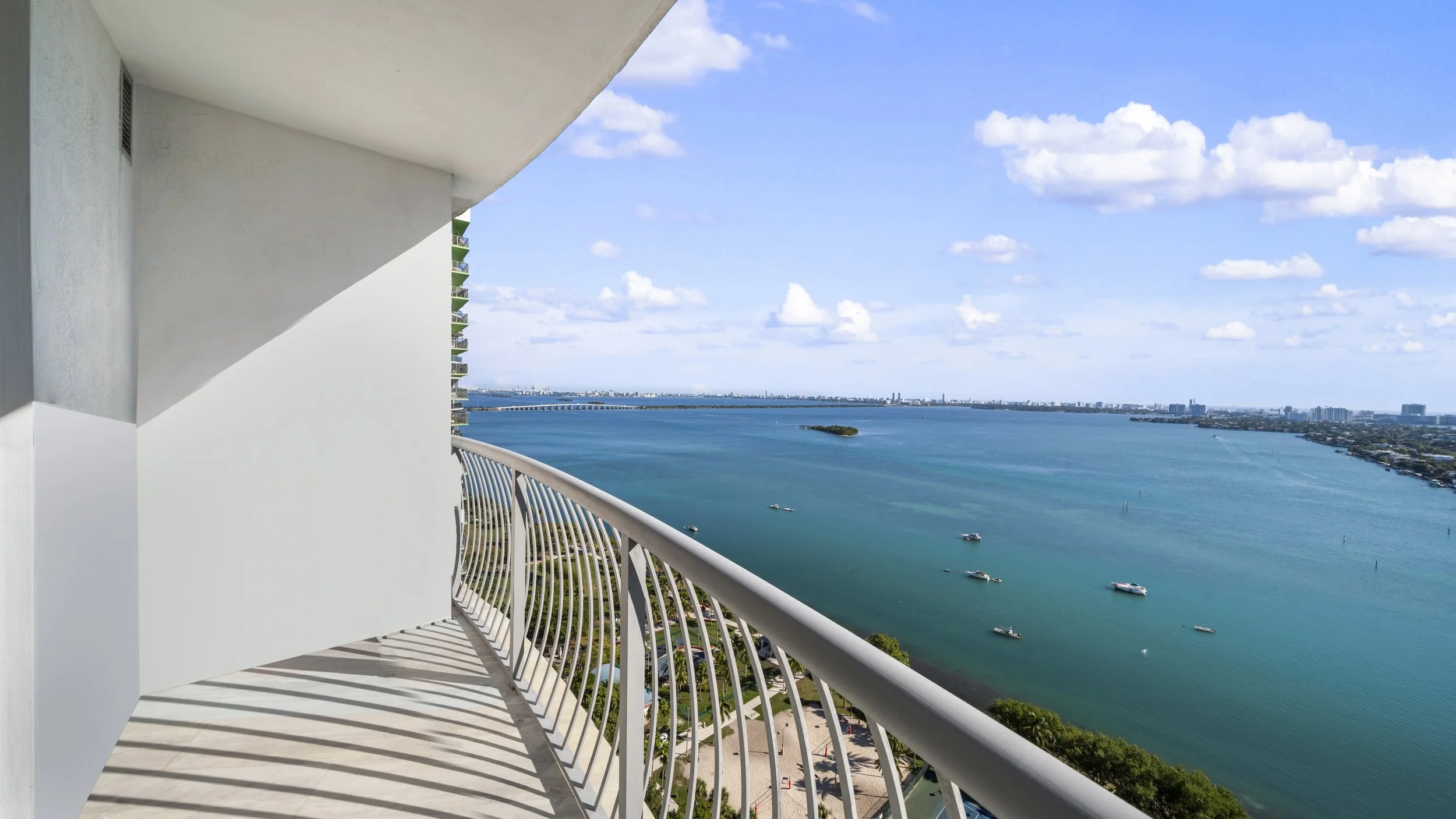 View from a high-rise balcony overlooking a large body of water with boats, a small island, and a city skyline in the distance on a partly cloudy day.