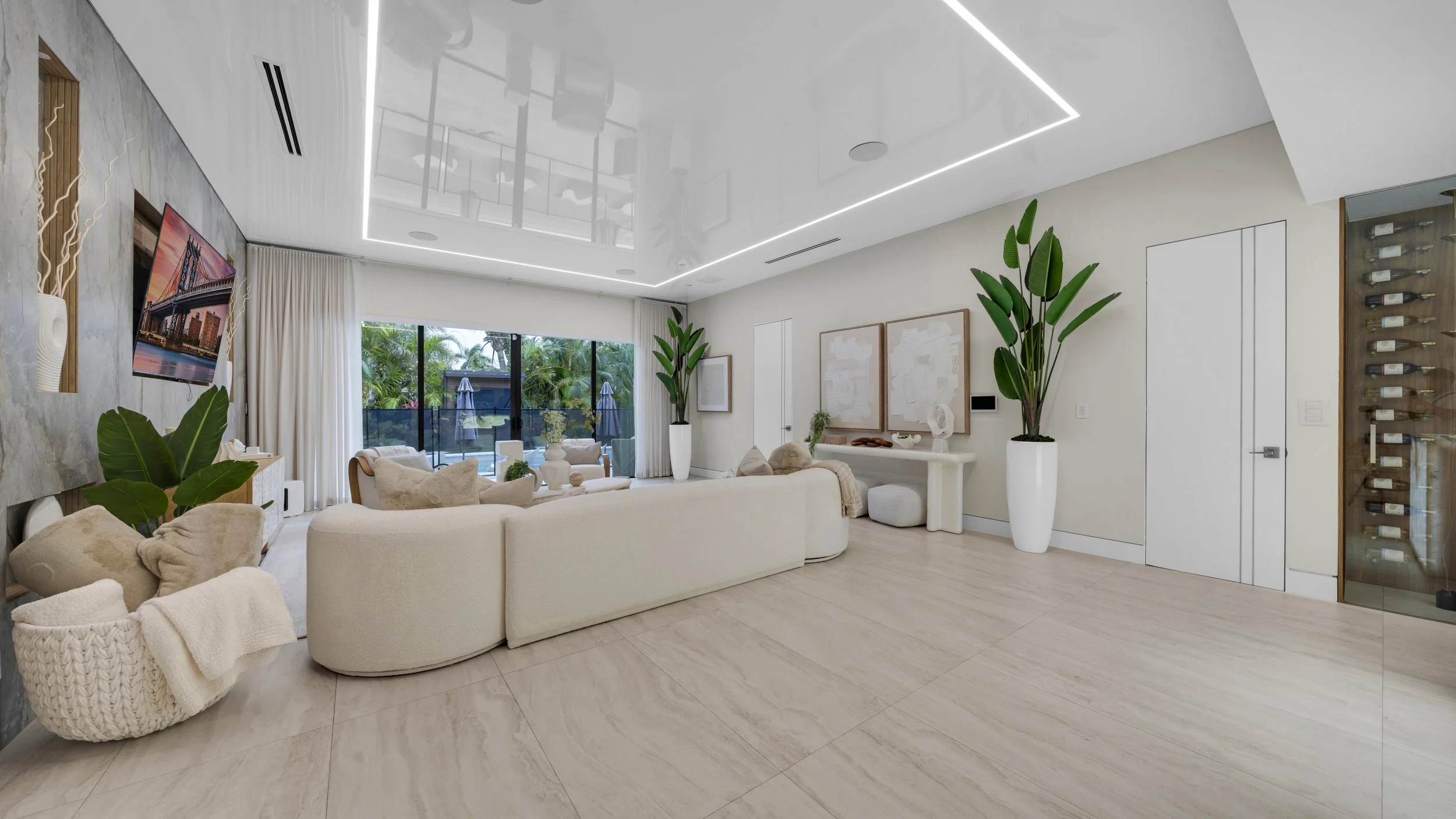 Modern living room with white sofas, large potted plants, artwork on the wall, a flat-screen TV, and sliding glass doors leading to a patio with outdoor furniture and umbrellas.