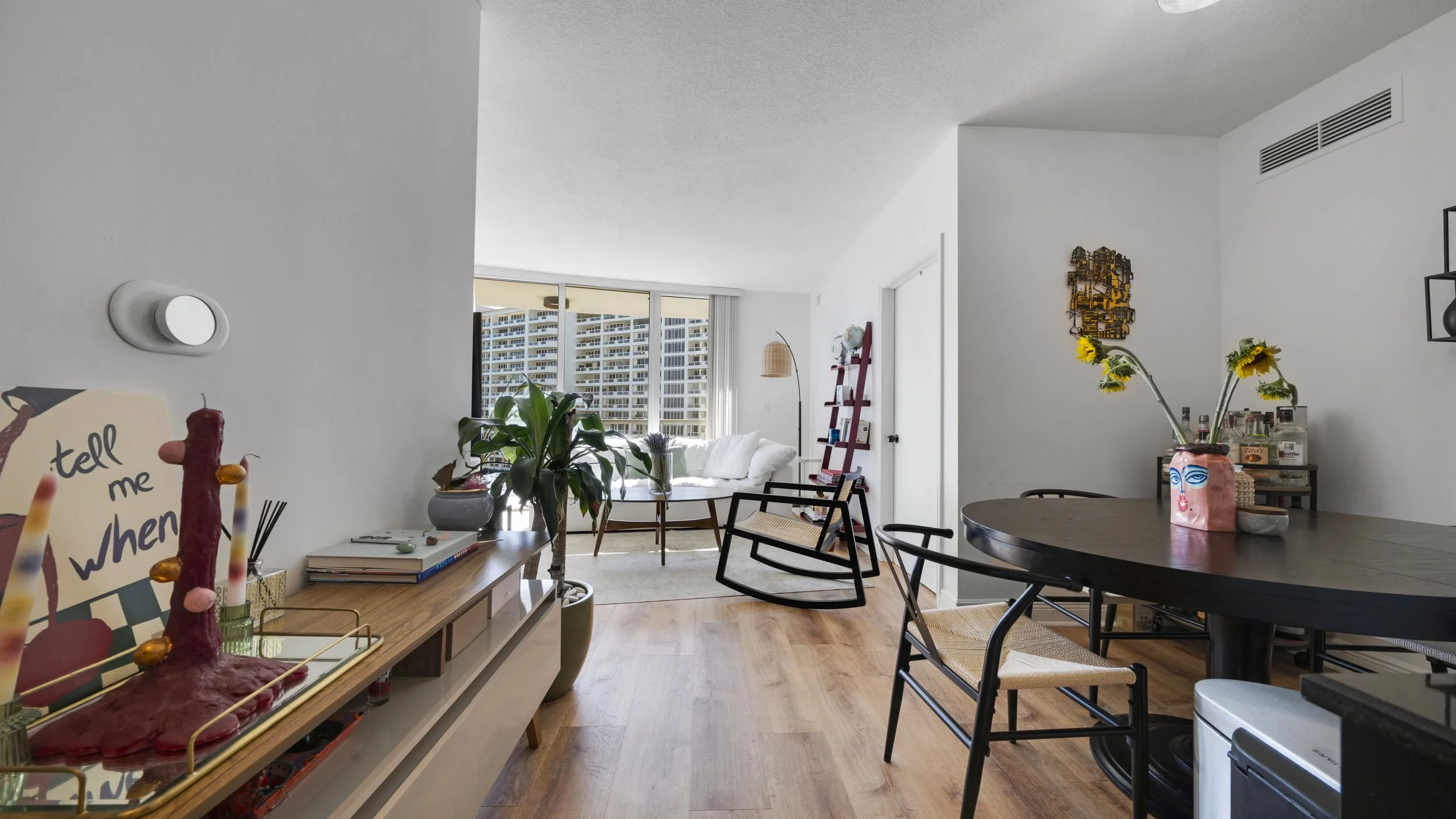 Living room with large window, white sofa, dining table with chairs, potted plants, and artwork on the walls.