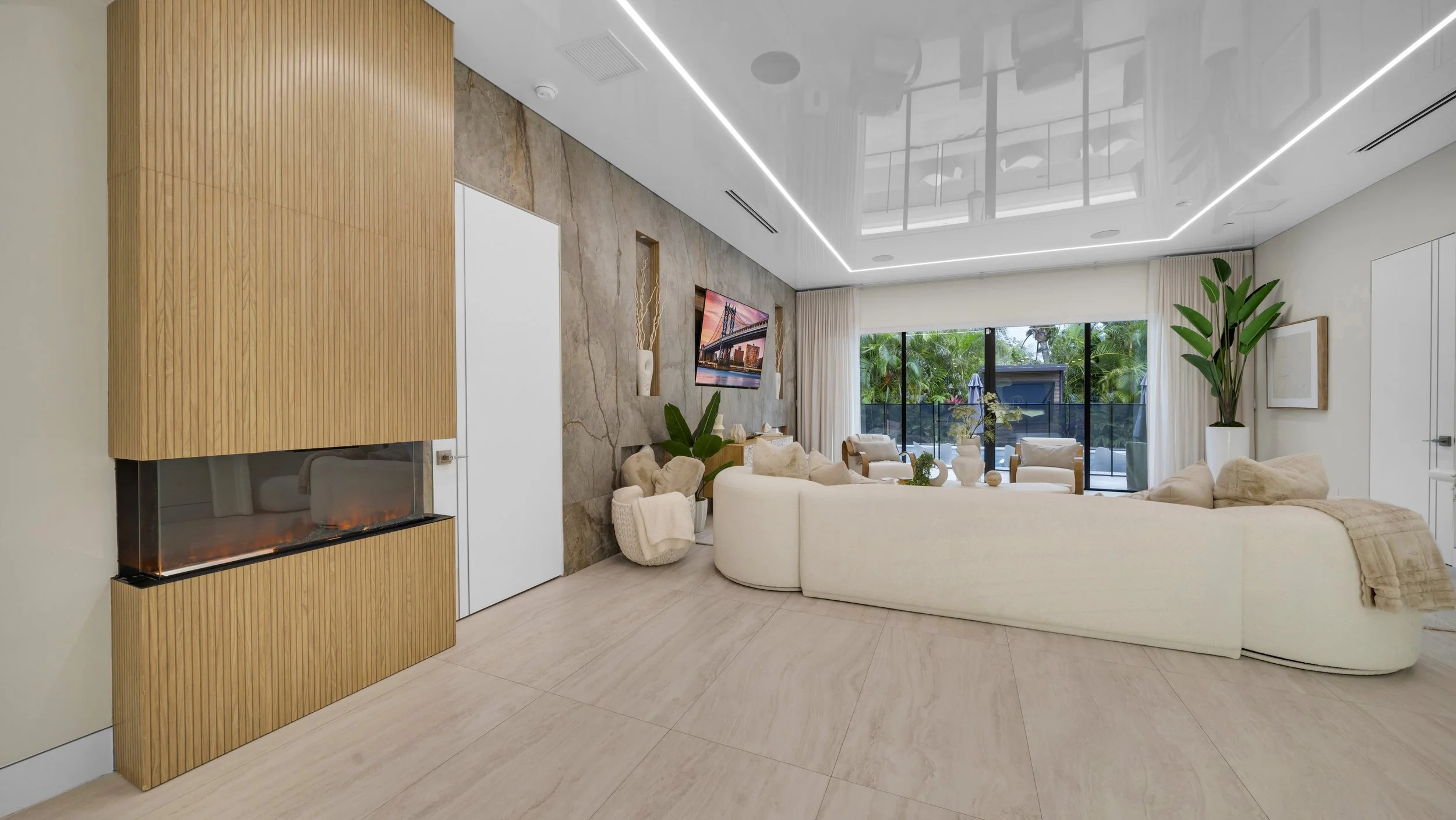 Modern living room with cream-colored sofa, large window with curtains, potted plants, and a flat-screen TV mounted on a textured wall.