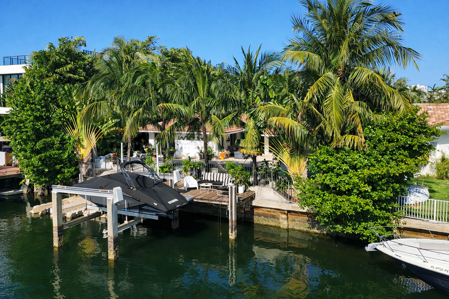 A lush tropical backyard with palm trees, patio furniture, and a dock with a jet ski and boats on calm water.