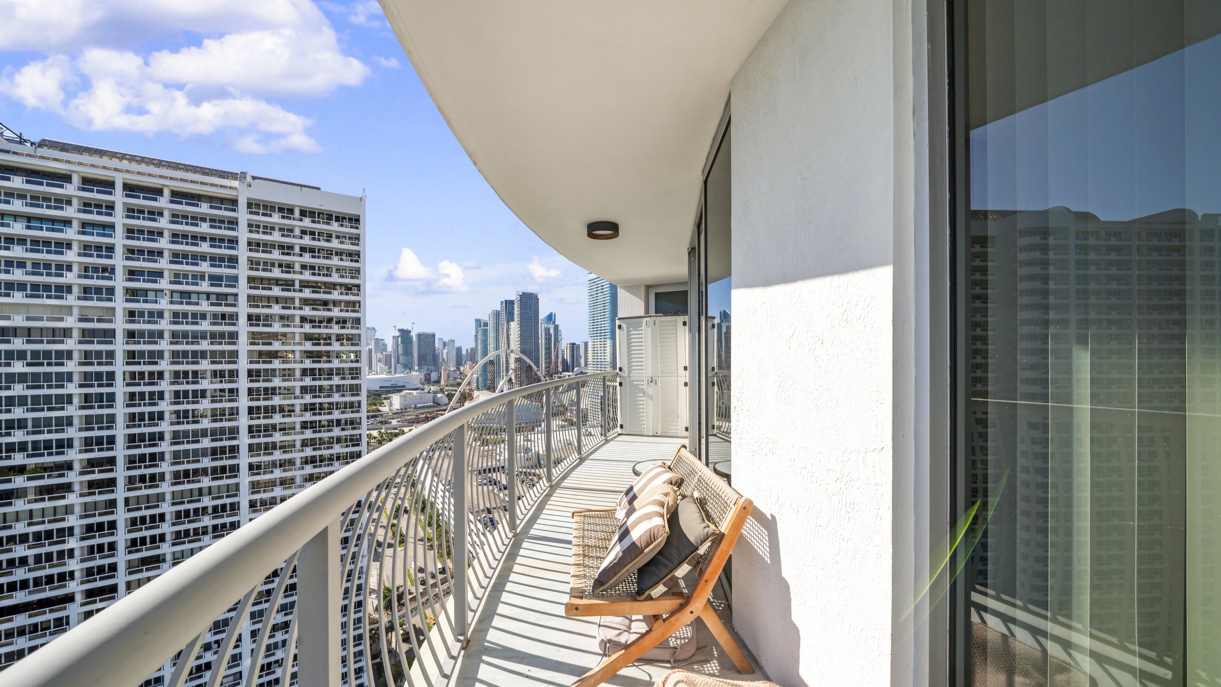 Balcony with chairs and pillows overlooking a city skyline with tall buildings and a blue sky.