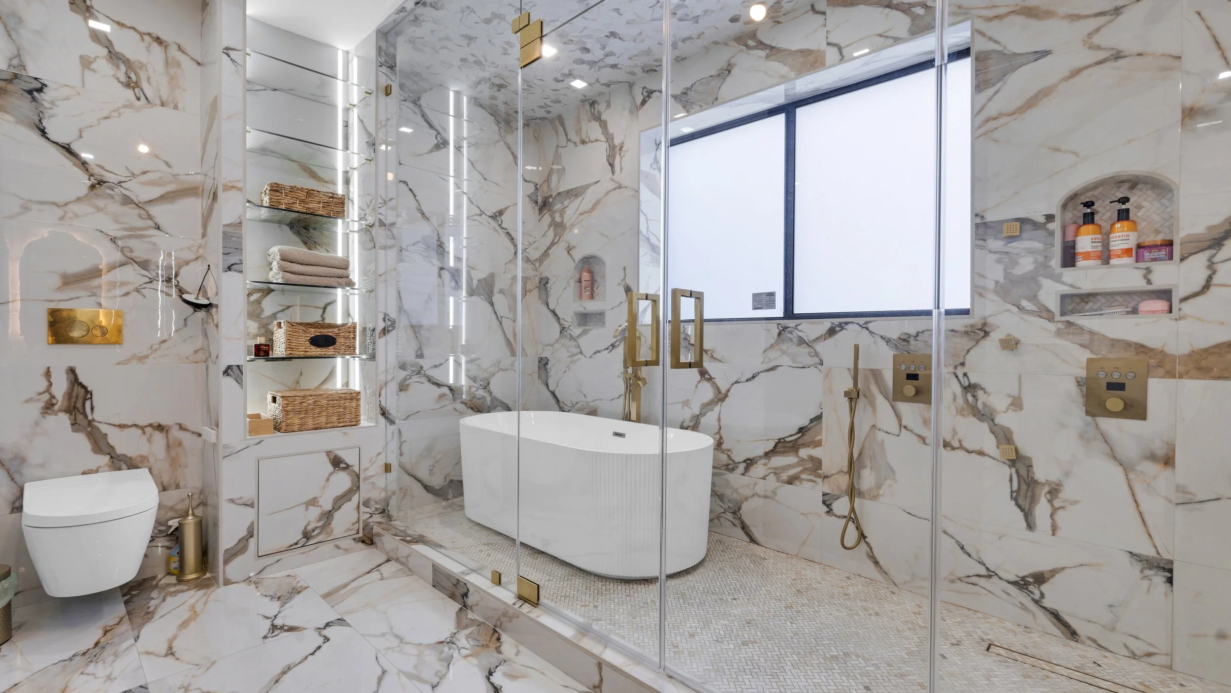 Luxurious bathroom with marble walls and floor, glass shower enclosure, and a freestanding white bathtub under a large window. Open shelving with towels, woven baskets, and toiletries. Modern fixtures with gold accents.