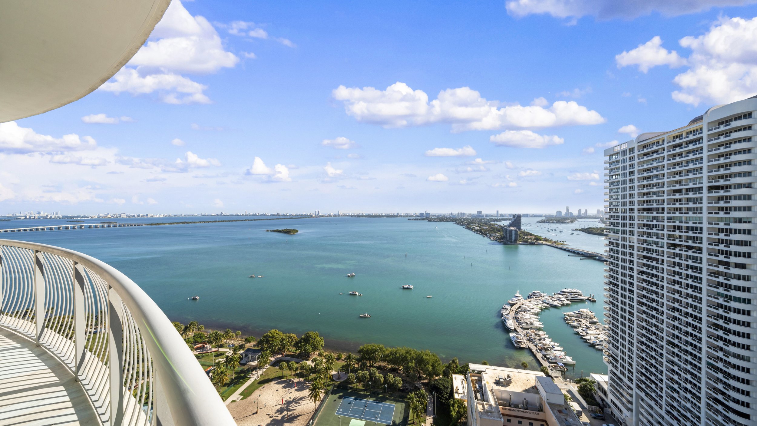 View from a high-rise balcony overlooking a large marina with boats, a stretch of water, and a cityscape in the distance under a partly cloudy sky.