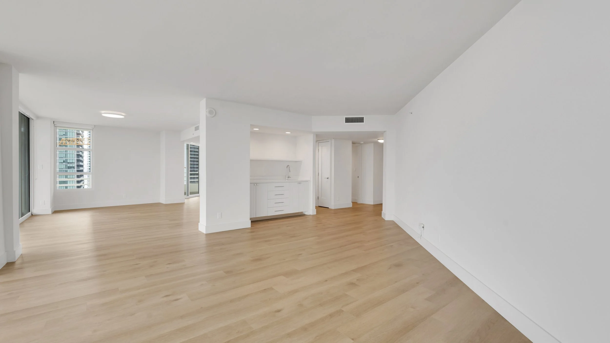 Empty, modern apartment living room with light wood flooring, white walls, large windows, and a small kitchenette with white cabinets.