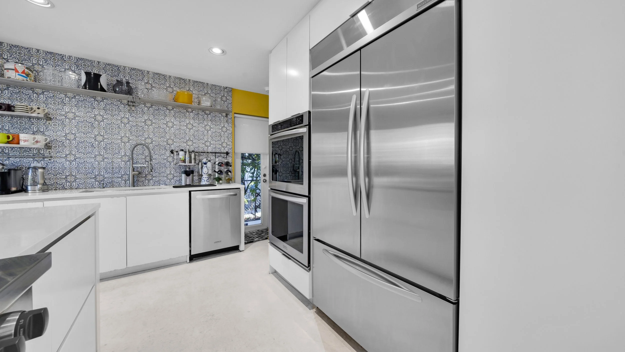 Modern kitchen with white cabinets, stainless steel refrigerator, double oven, patterned tile backsplash, and small door leading outside.