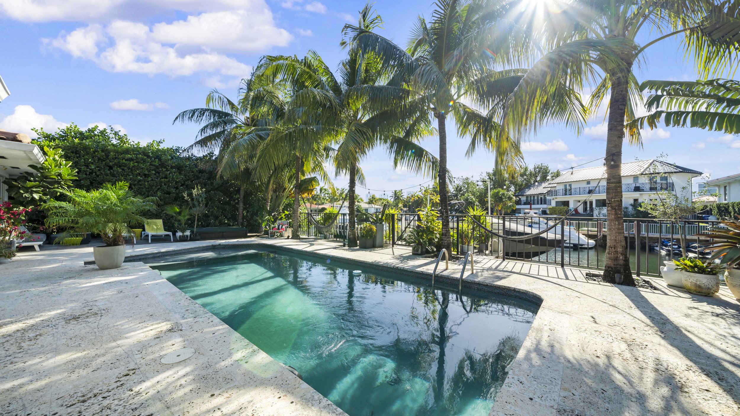 Backyard with swimming pool, palm trees, lounge chairs, and a canal view with boats and houses under a partly cloudy sky.