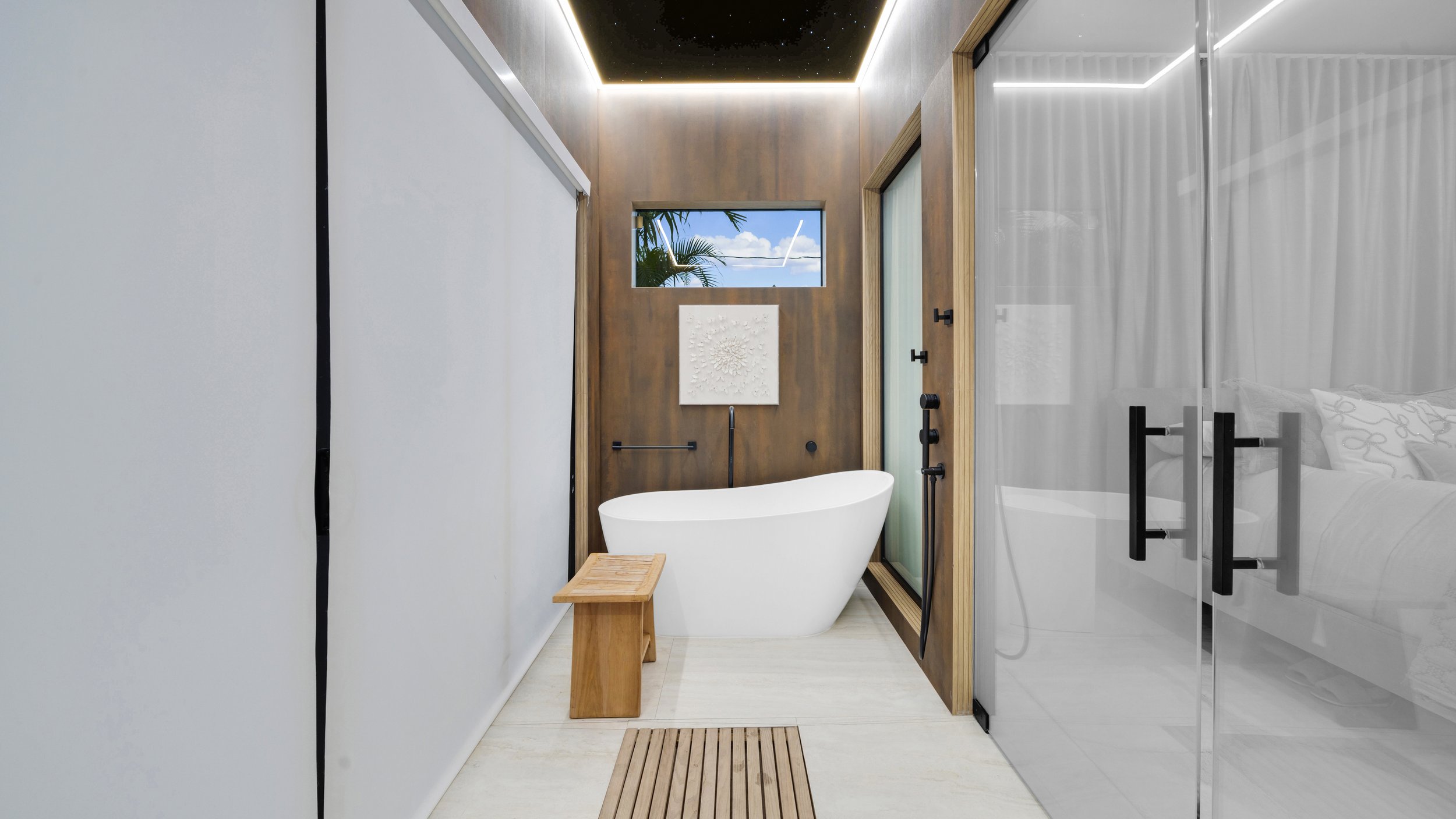 Modern bathroom with a standalone bathtub, wooden bench, and a small window showing trees and sky.