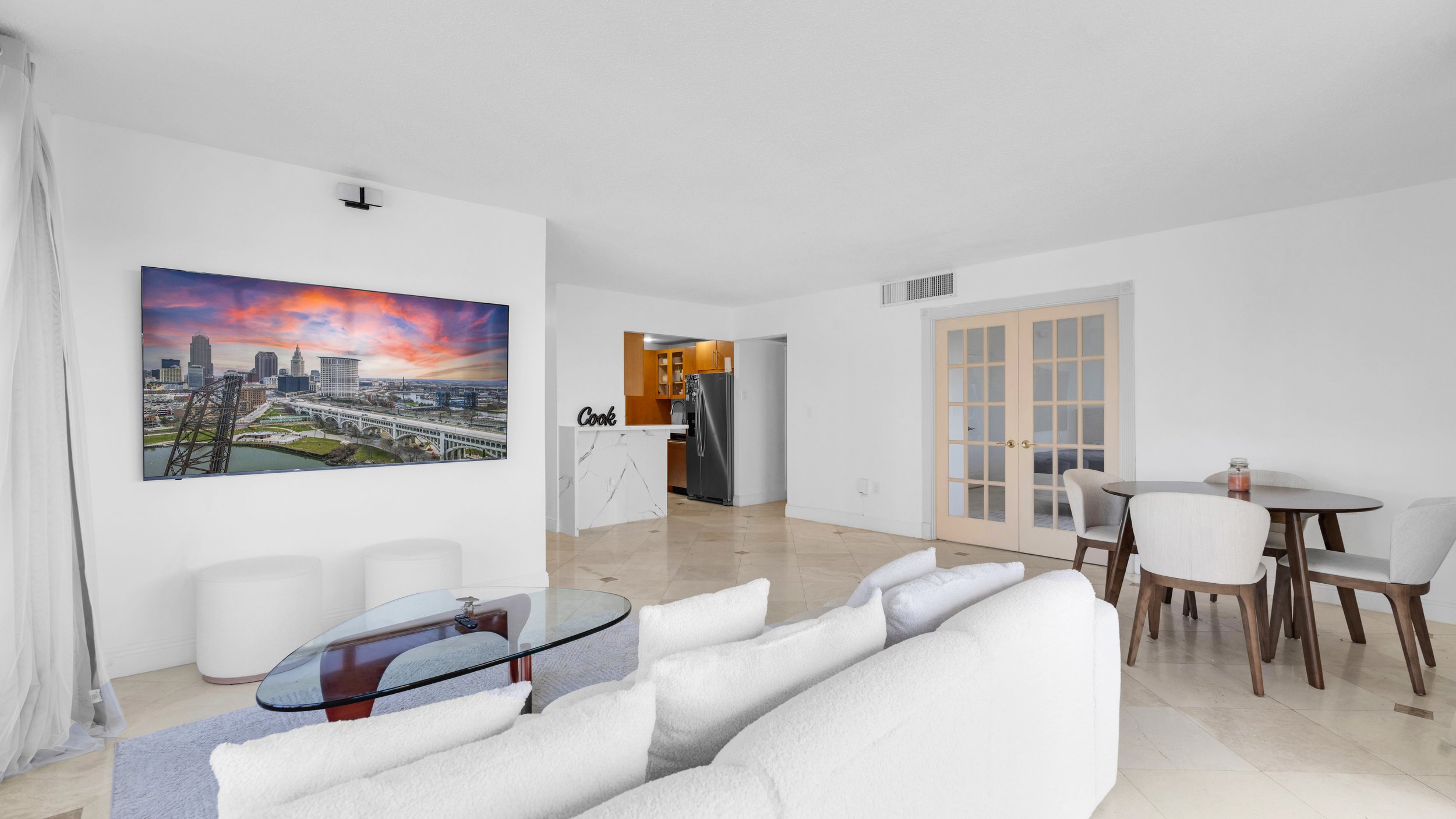 Living room with white sofa, glass coffee table, circular white ottomans, dining table with four chairs, French doors, and a large wall-mounted TV showing a cityscape at sunset.