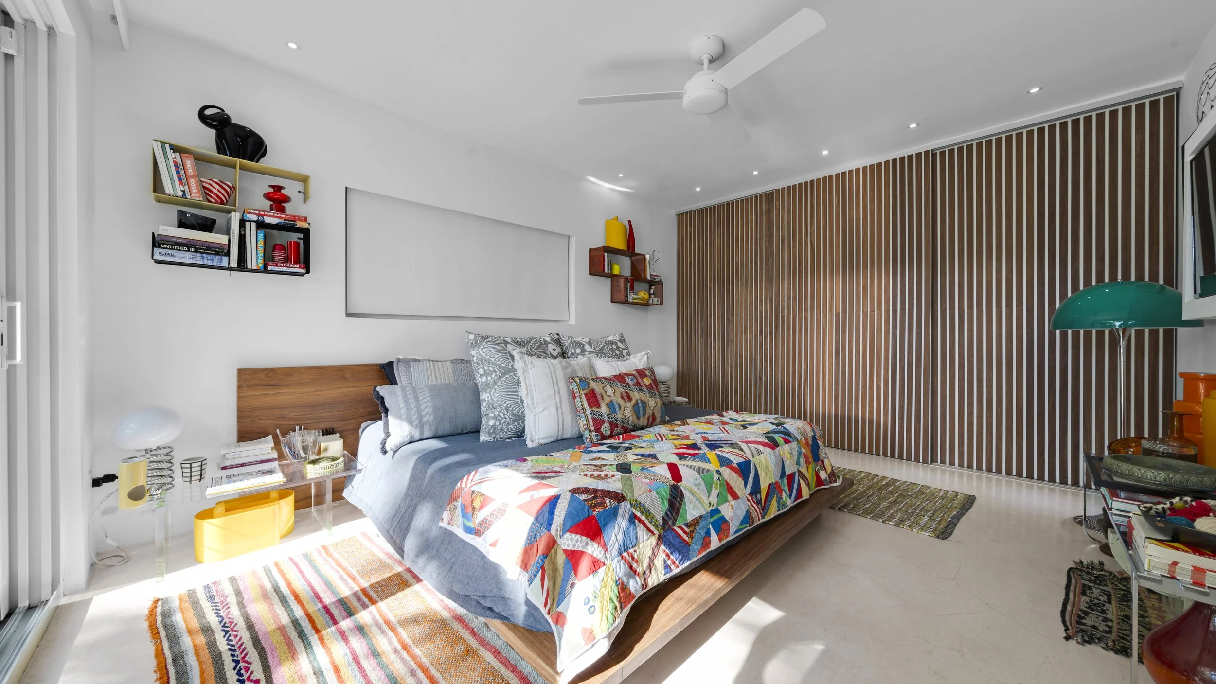 A modern bedroom with a wooden bed, colorful quilt, and multiple pillows. The room features a wooden paneled wall, white walls, a ceiling fan, and various shelves with books and decorative items. There is a glass side table with books and lamps, and 
