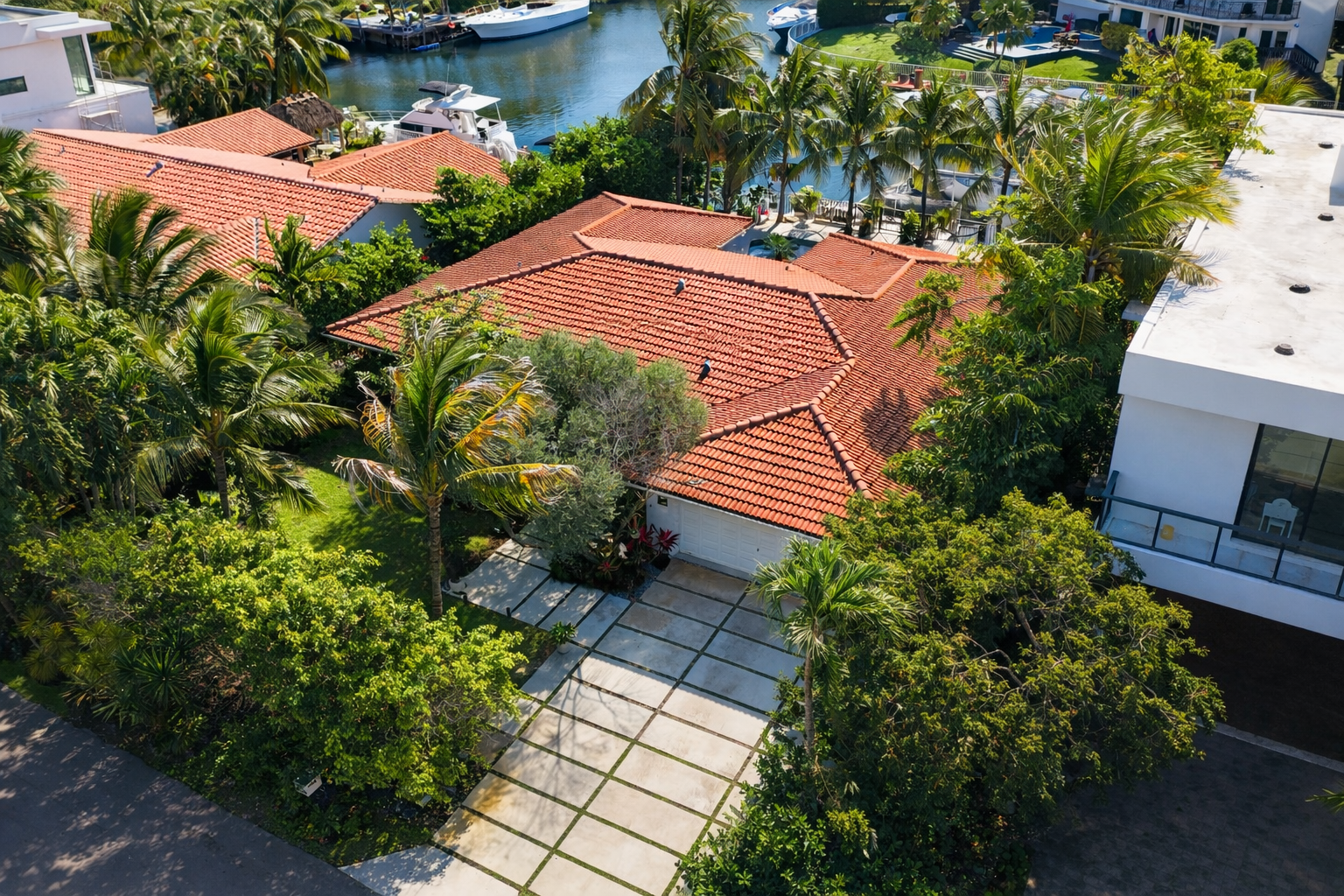 An aerial view of a tropical residential area with a house featuring a red-tile roof, surrounded by lush green trees and plants, with a paved driveway leading to a white garage door. Nearby is a modern white building with a balcony, and in the backgr