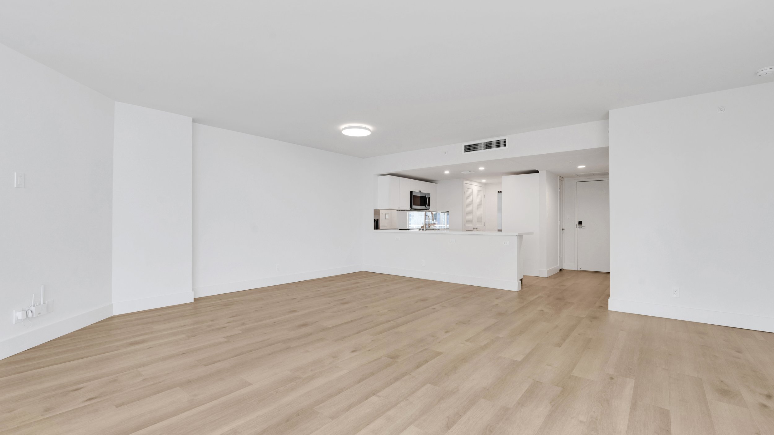 Empty living room with white walls, light wood flooring, and an open kitchen area in the background.