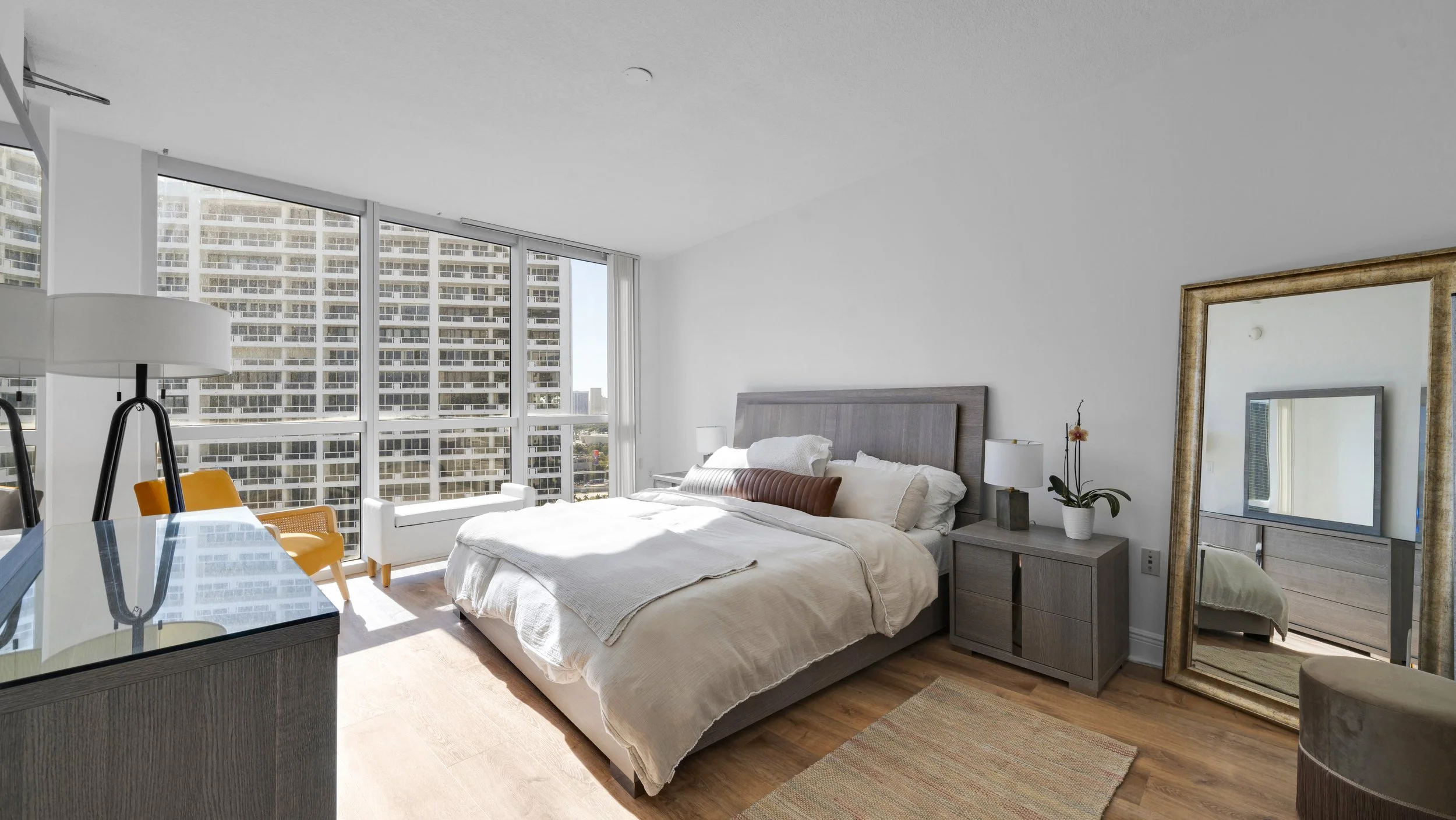 A modern bedroom with a large bed, nightstands with lamps, floor-to-ceiling windows showing city buildings, a large standing mirror, a sitting area outside the window, and wooden flooring.