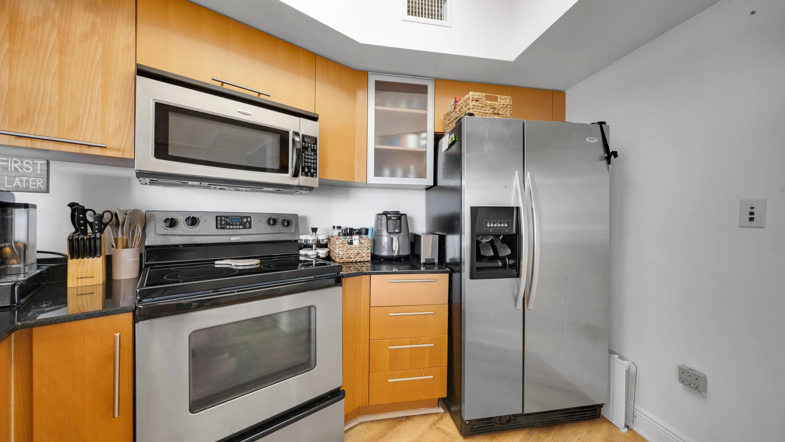 Modern kitchen with wooden cabinets, stainless steel refrigerator, black countertop, microwave oven, stove, and small appliances.