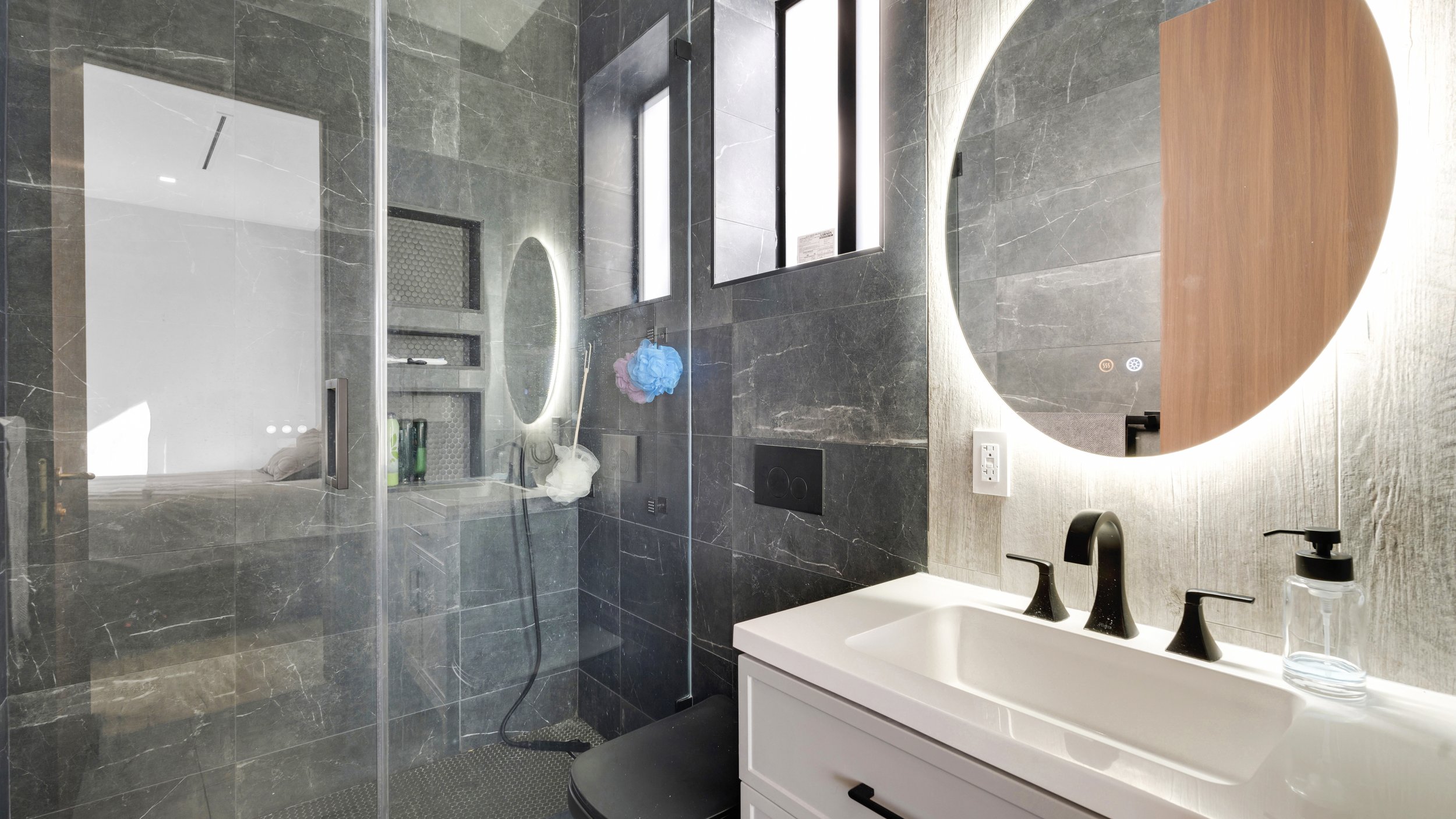 Modern bathroom with dark gray marble tiles, a white sink with black fixtures, a round illuminated mirror, and a glass-enclosed shower area with toiletries and cleaning items inside.