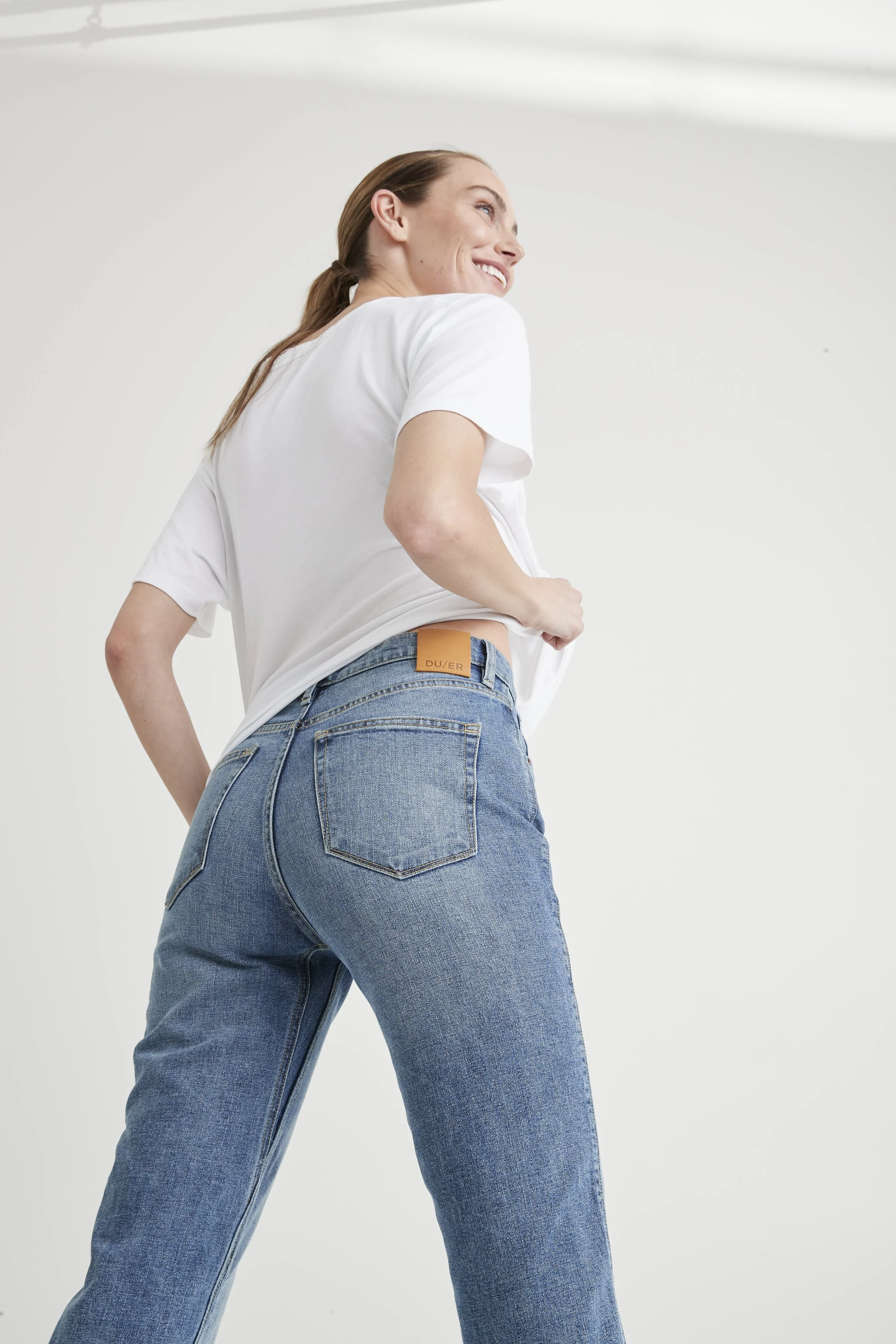 A woman with long brown hair, wearing a white t-shirt and blue jeans, is smiling and looking upward. She is standing against a plain white background.