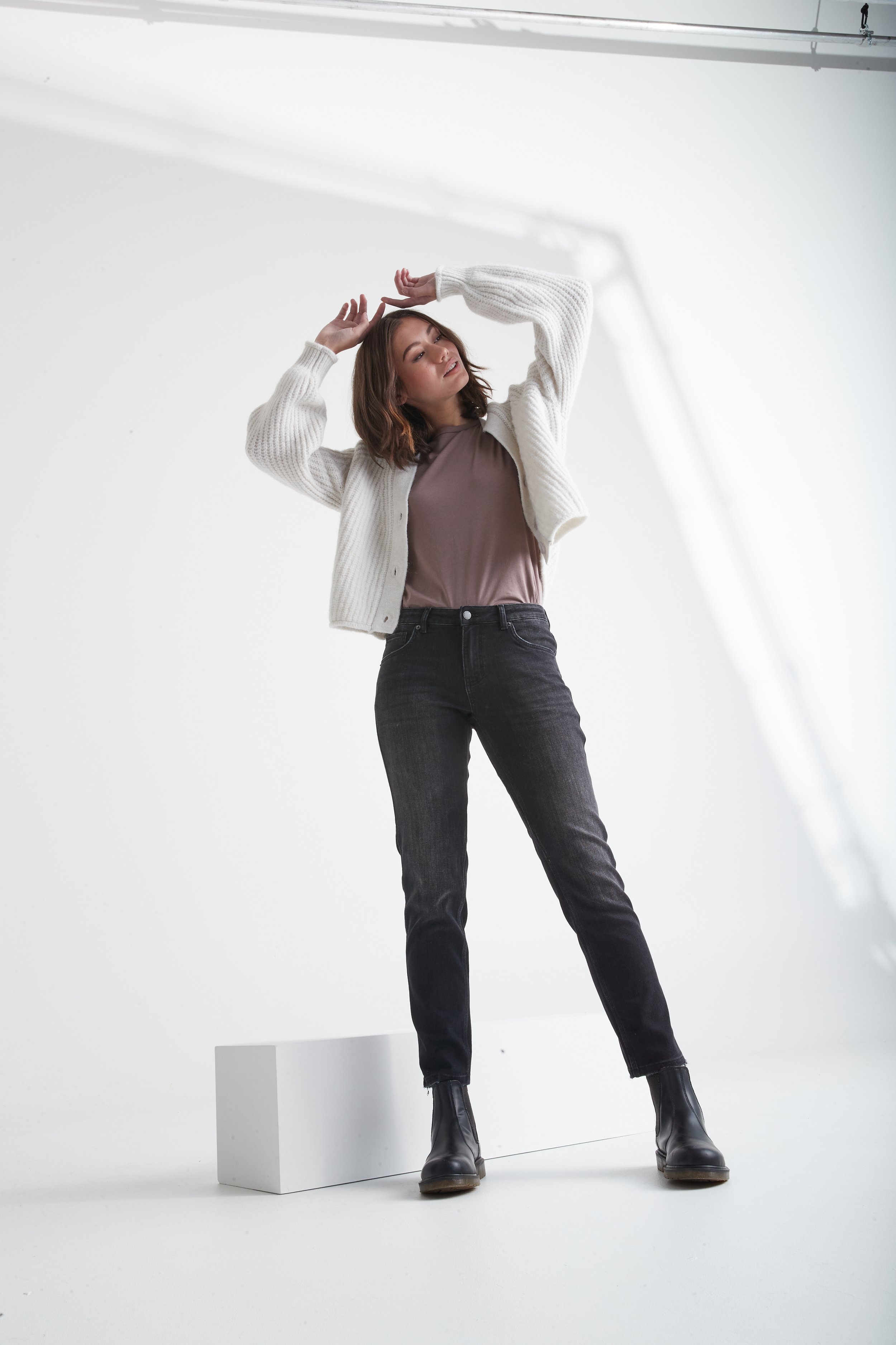 A woman standing on a white platform in a photography studio with a plain white background, wearing a beige top, white cardigan, black jeans, and black boots, posing with her hands above her head.