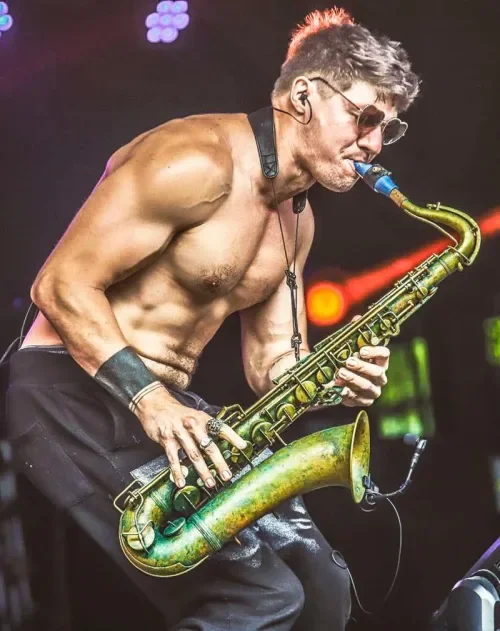 A shirtless man wearing sunglasses and wristbands playing a saxophone on stage, with stage lights in the background.
