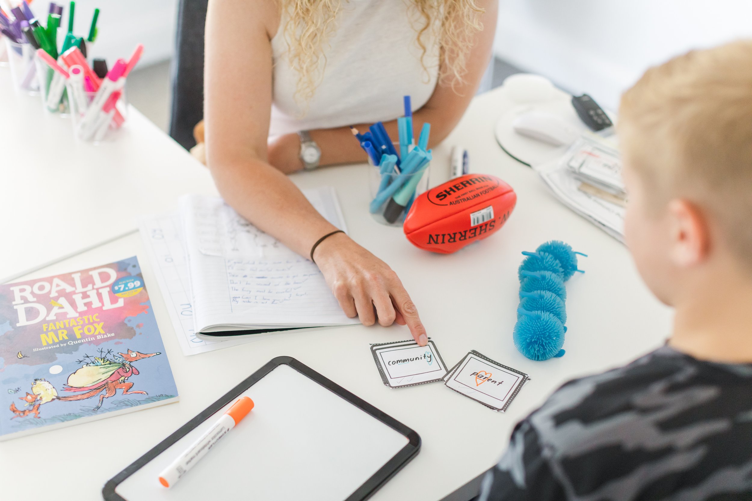 A woman is sitting at a white desk, pointing at a card with the word 'community' written on it, while a young boy sits across from her. The desk has various items including a book titled 'Roald Dahl Fantastic Mr. Fox,' a whiteboard marker, a tablet, and several other cards with words like 'parent' and 'community.' There are also stationery items like pens in cups, a small inflatable Australian football, and a blue plush toy resembling a caterpillar.
