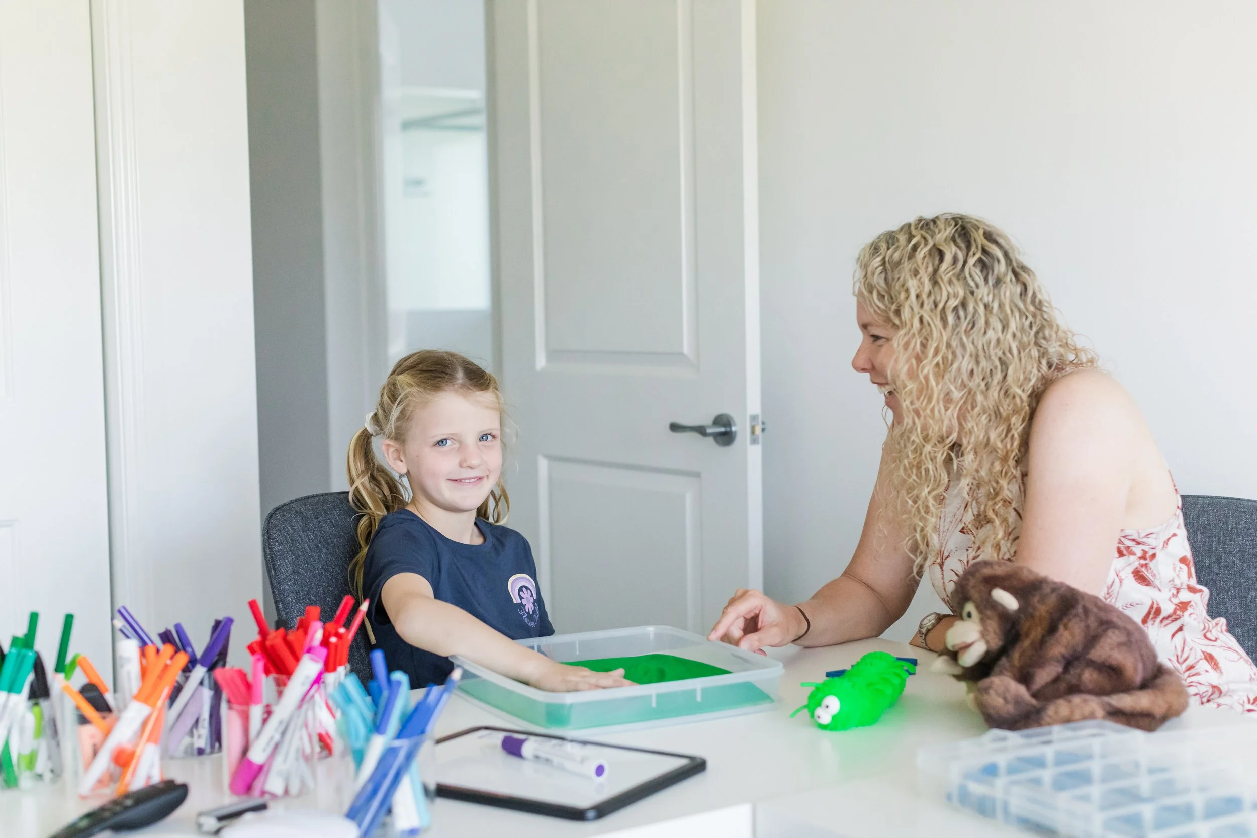 A woman and a young girl seated at a table with toys and art supplies. The girl has her hand in a green slime box and the woman is smiling. There are plush toys and colored markers on the table.