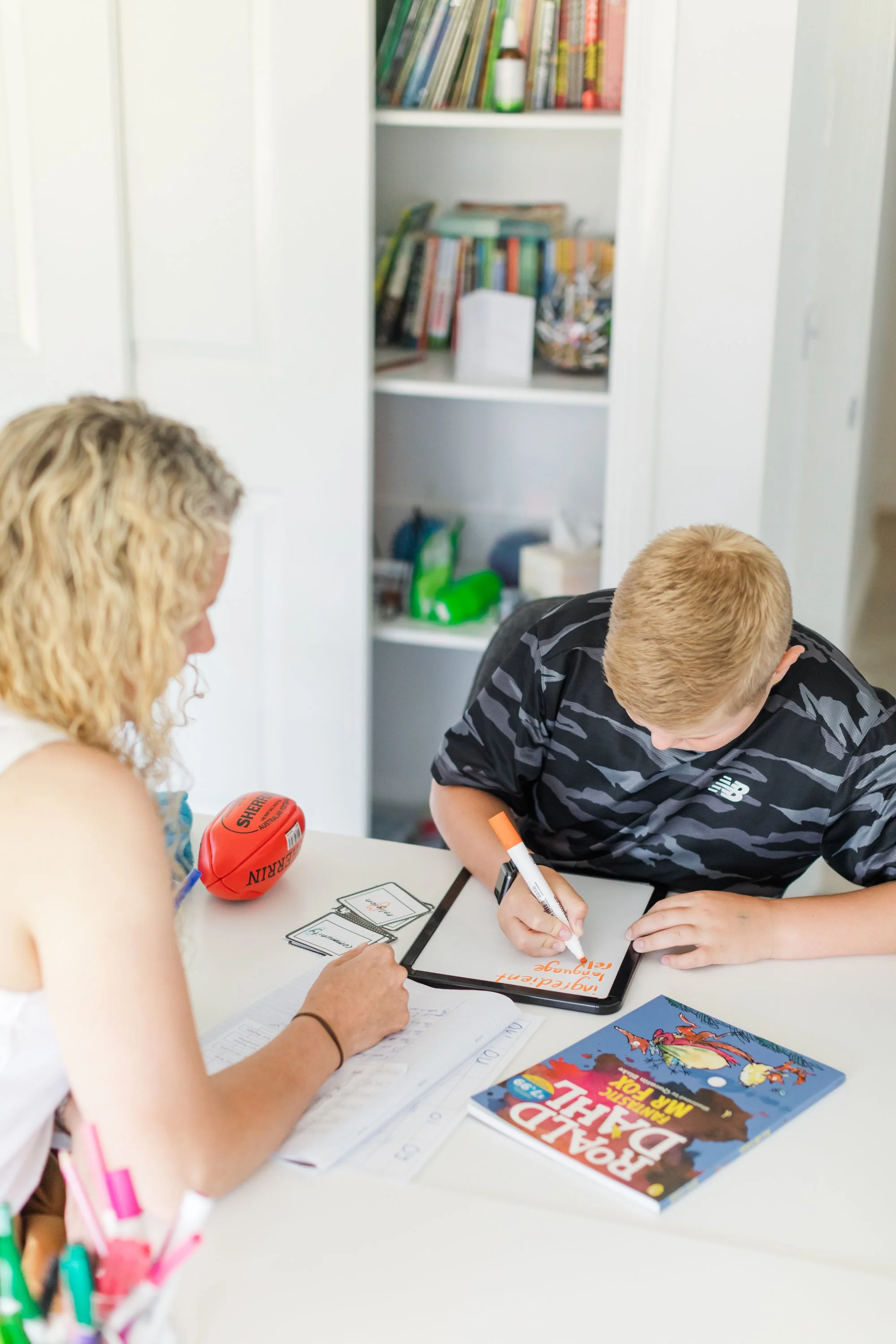 A woman and a young boy sitting at a table, with the woman helping the boy with homework. There is a book titled 'Rainbow Dale, The Legend' on the table, along with a small red football, markers, and a notebook.