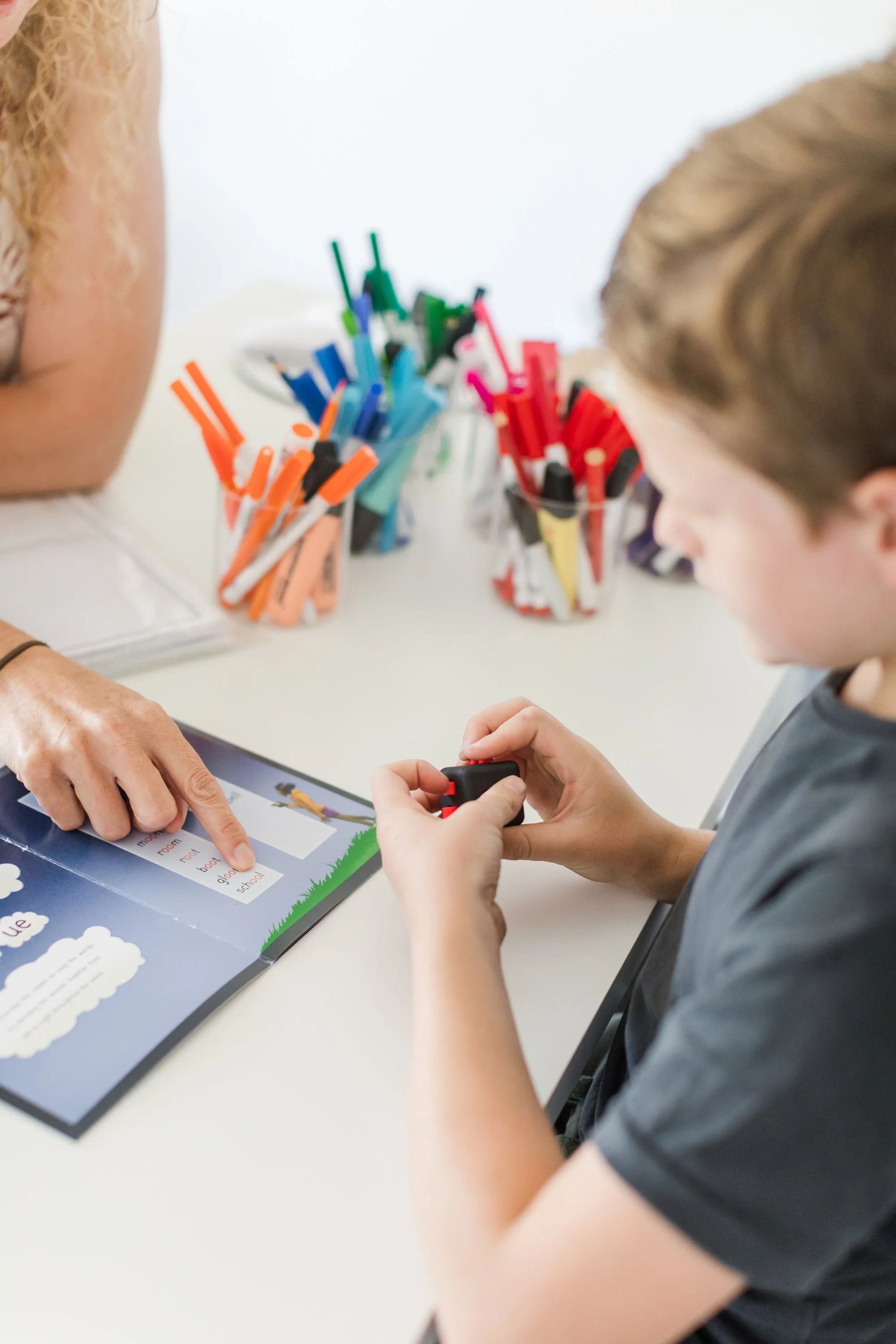 Child and adult looking at a colorful educational book on a white table with various colored markers in the background.