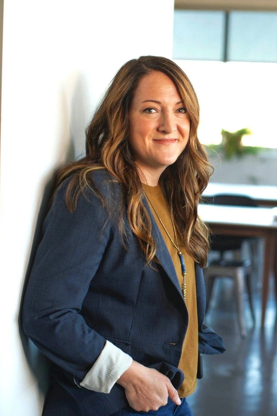 A woman with long, wavy brown hair, wearing a blue blazer and a mustard-coloured top, leaning against a white wall in a classroom setting.