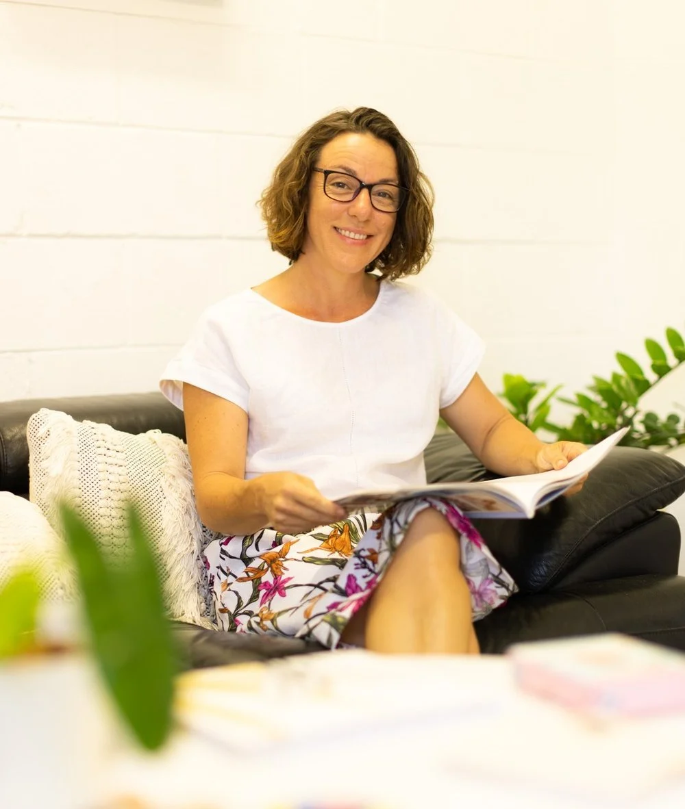 A woman with glasses and curly brown hair sitting on a black leather couch, smiling, reading a magazine, with a white wall and green plants in the background.