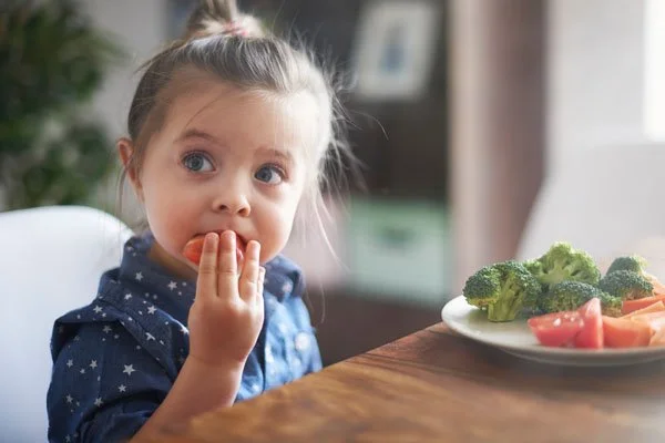 A young girl with light brown hair in a top knot, wearing a blue star-patterned shirt, sitting at a table with a plate of broccoli and sliced tomatoes, eating a snack.