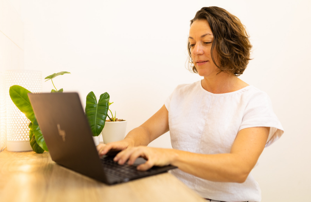 Woman working on a laptop at a wooden desk with plants and a light behind her.