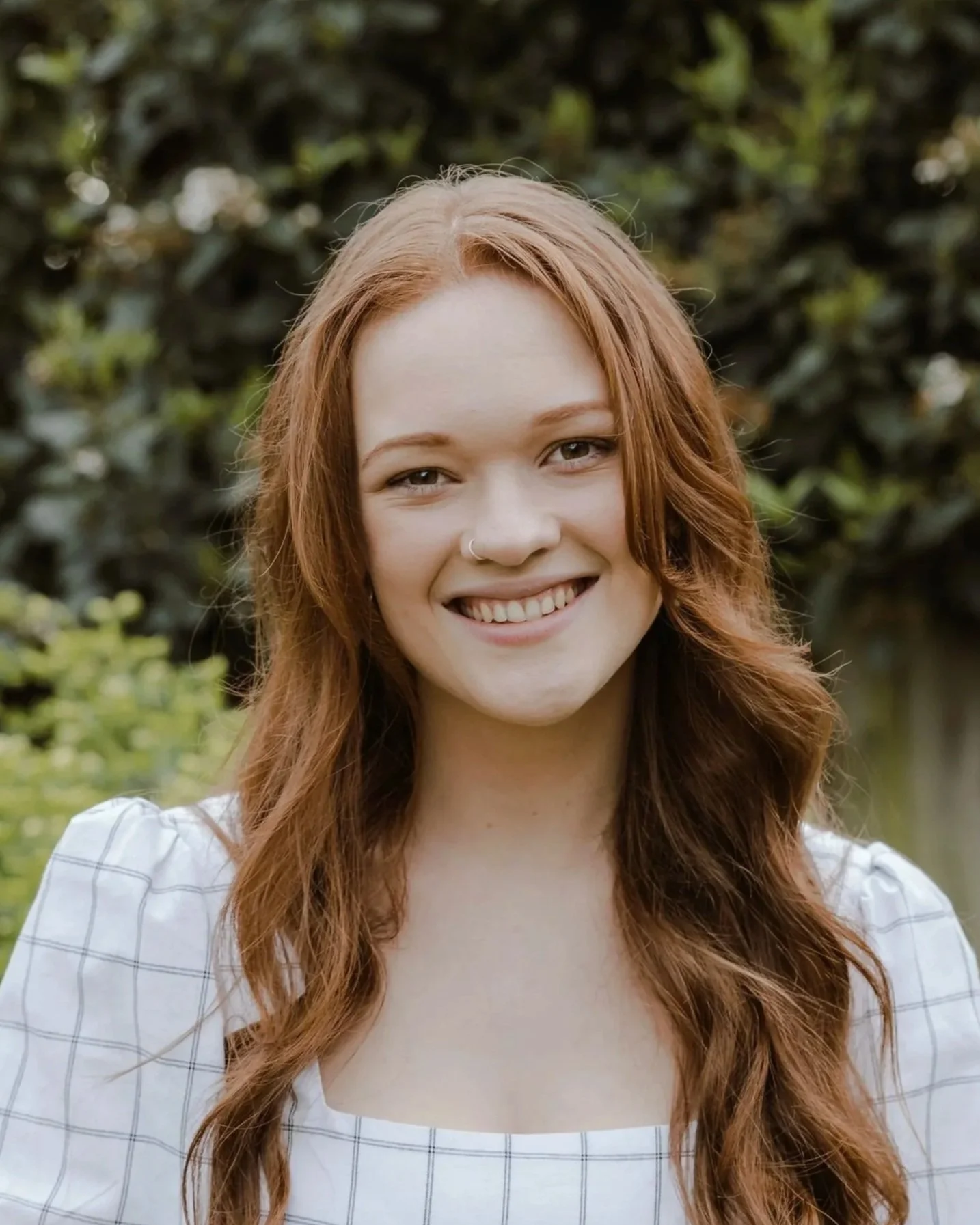 A young woman with long red hair smiling outdoors in front of greenery.
