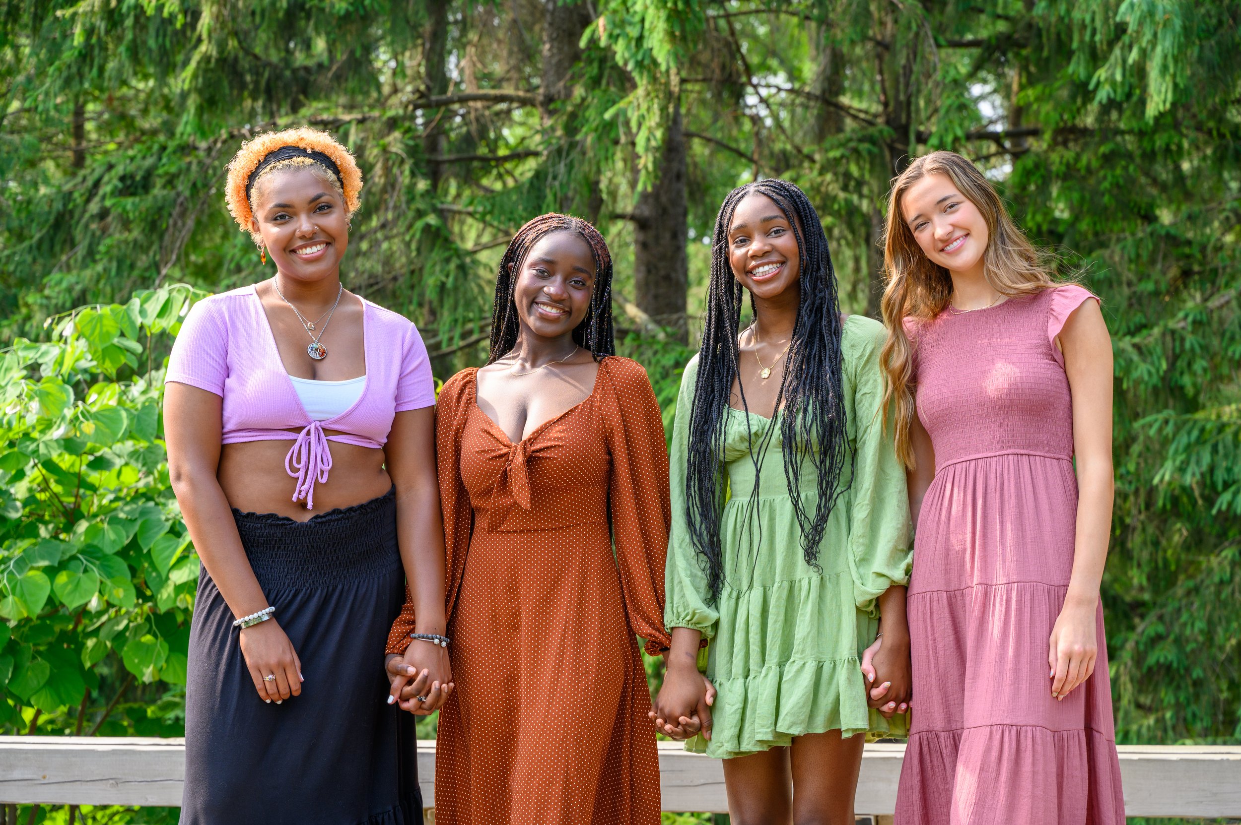 Four women standing outdoors, smiling and holding hands, with trees in the background.
