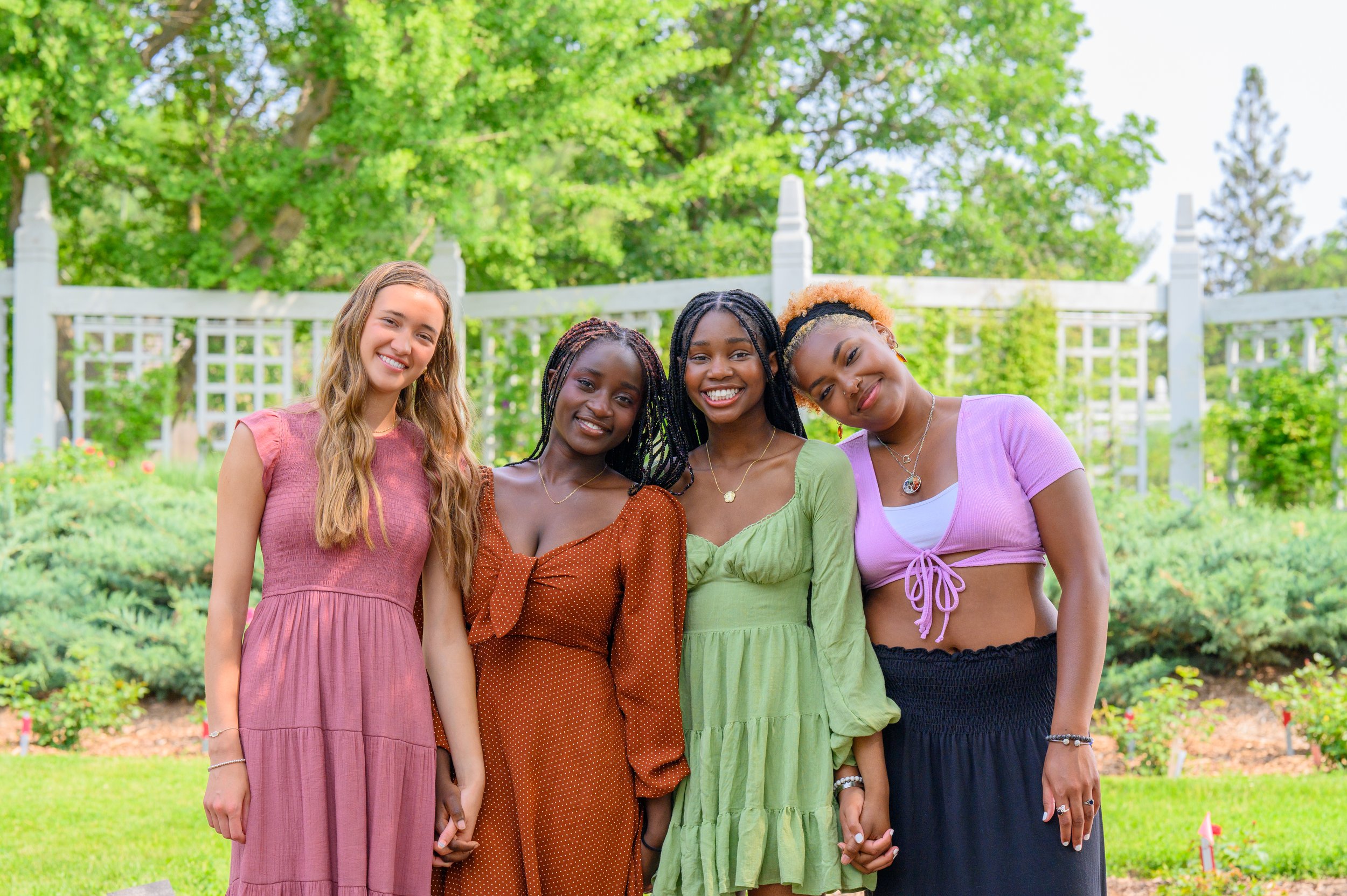Four diverse young women smiling and holding hands, standing outdoors in a garden with green trees and a white lattice fence in the background. They are wearing colorful dresses and casual tops.