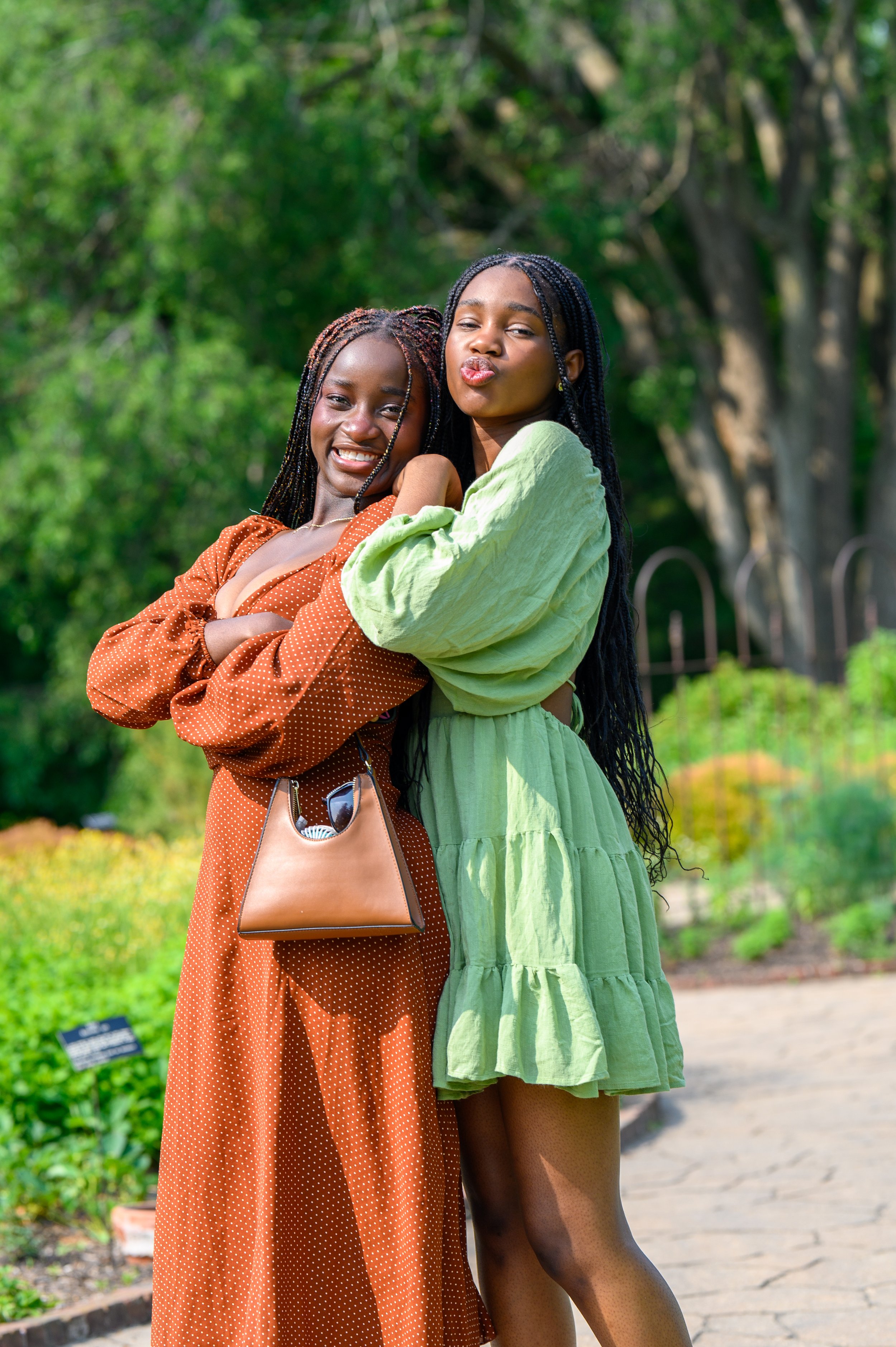 Two women with braided hair smiling in a garden, one in a brown dress with a handbag, the other in a green dress.