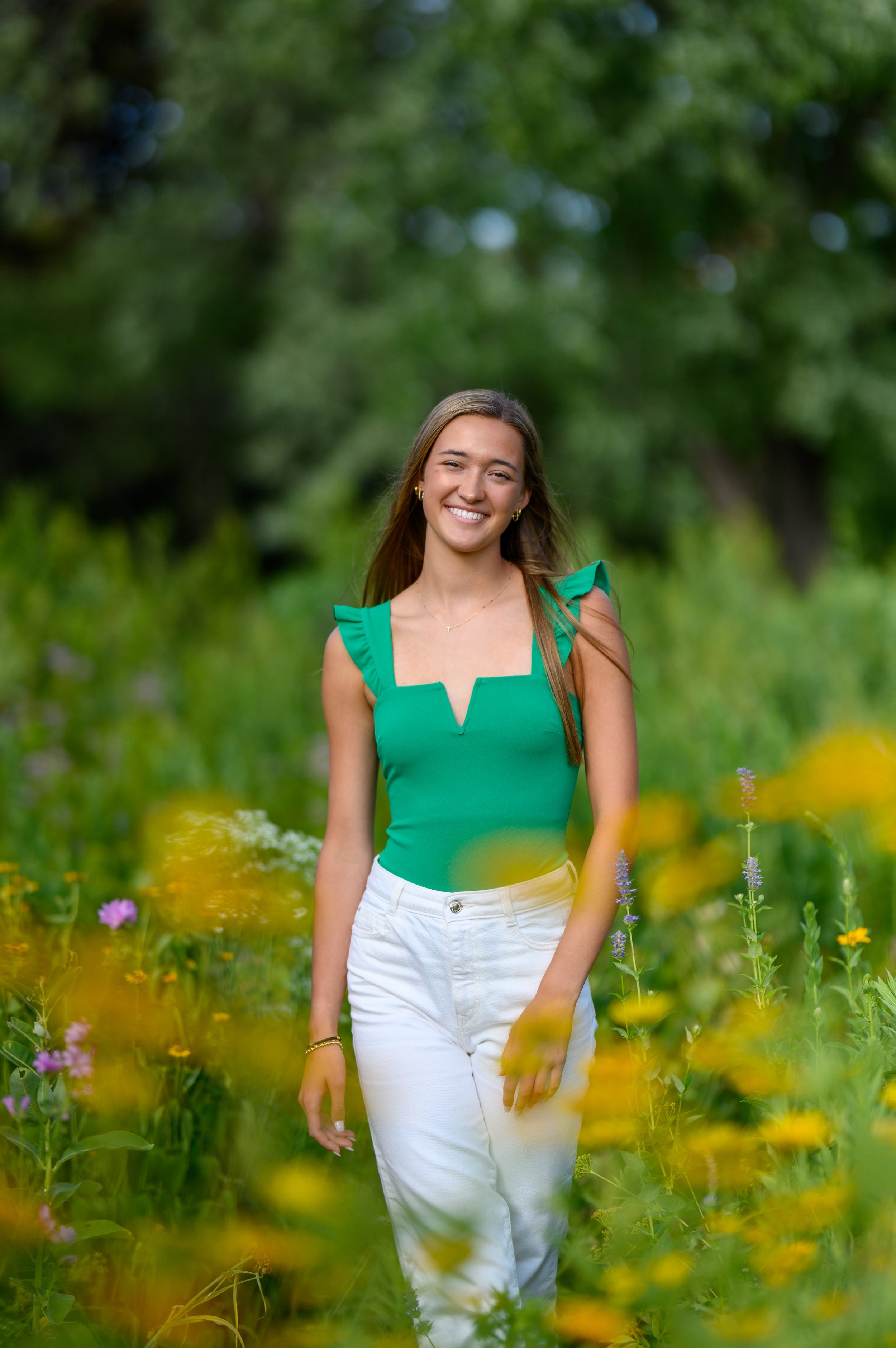 Woman in a green top and white pants standing in a flower garden with a blurred green and yellow background.