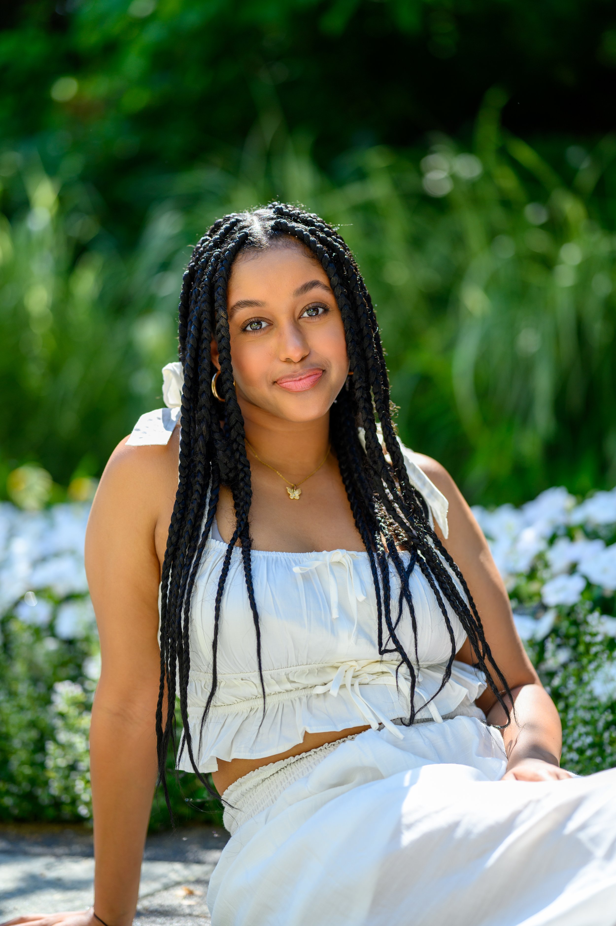 Woman with braided hair in white dress sitting outdoors with greenery and flowers.