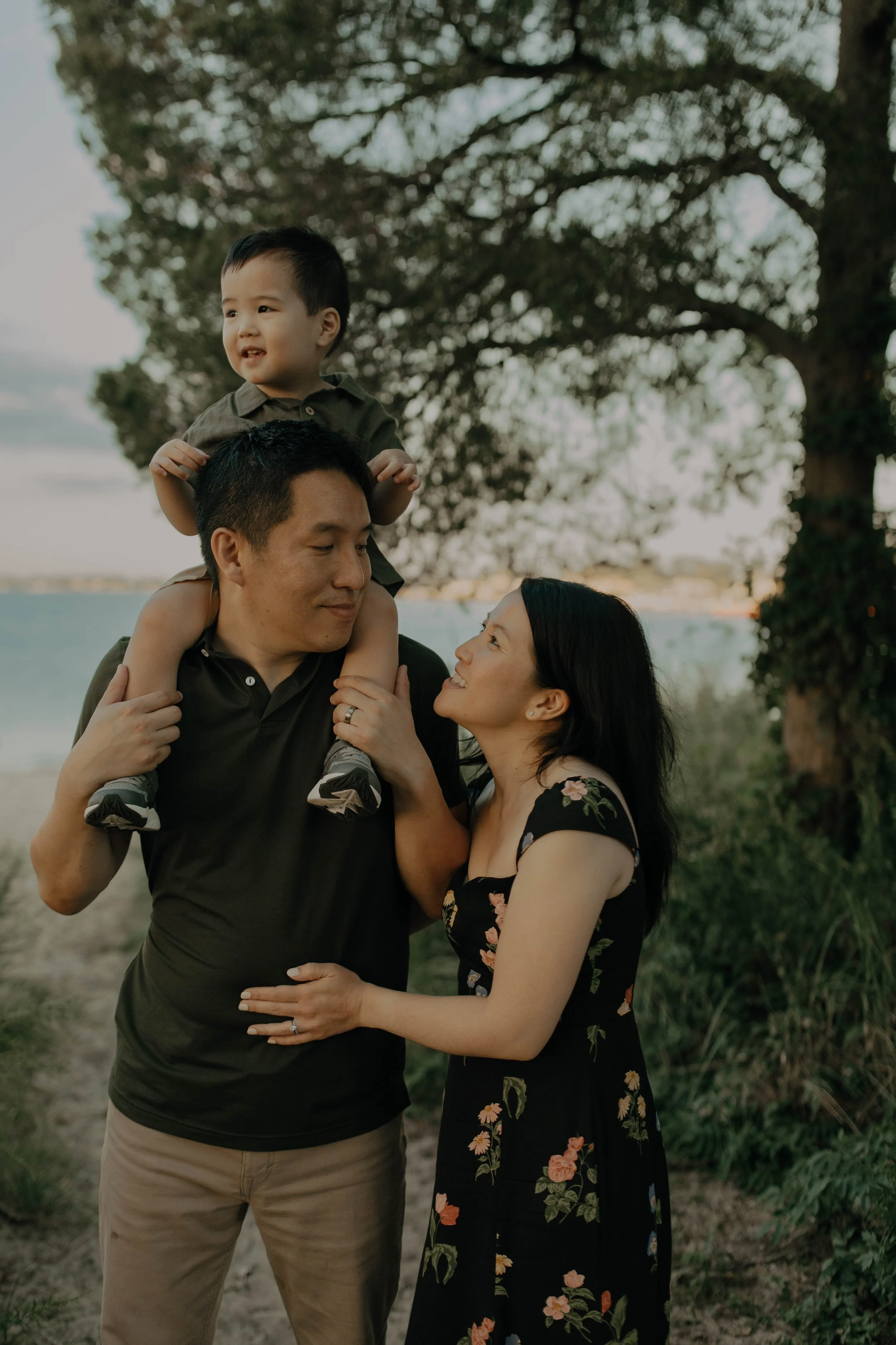 A family of three enjoying time outdoors at sunset. The father is carrying their young son on his shoulders, and the mother is holding the father's arm while gazing at him. There are trees and a body of water in the background.