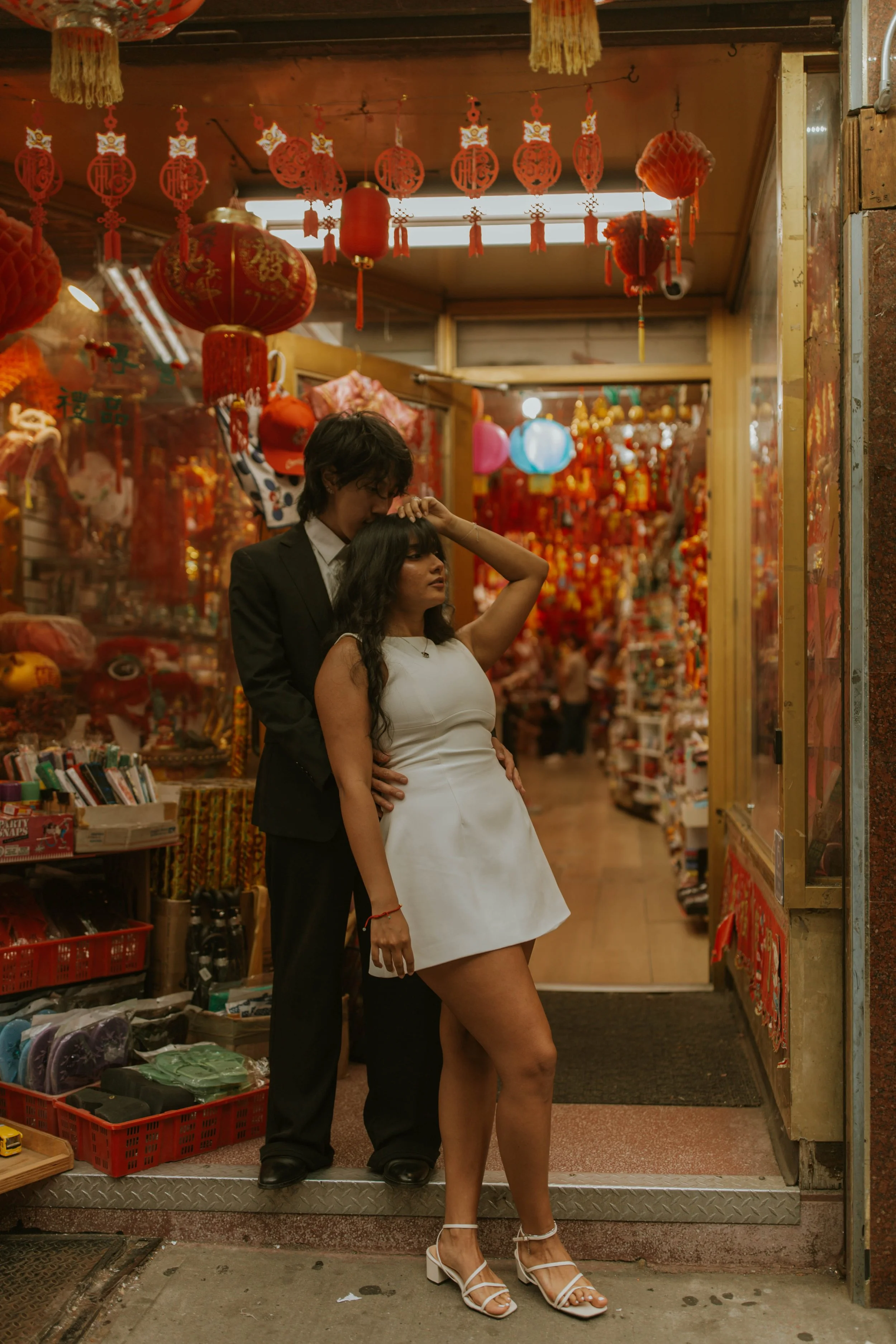 A woman in a white dress and white heels standing in front of a man in a black suit inside a shop decorated with red Chinese lanterns and decorations.