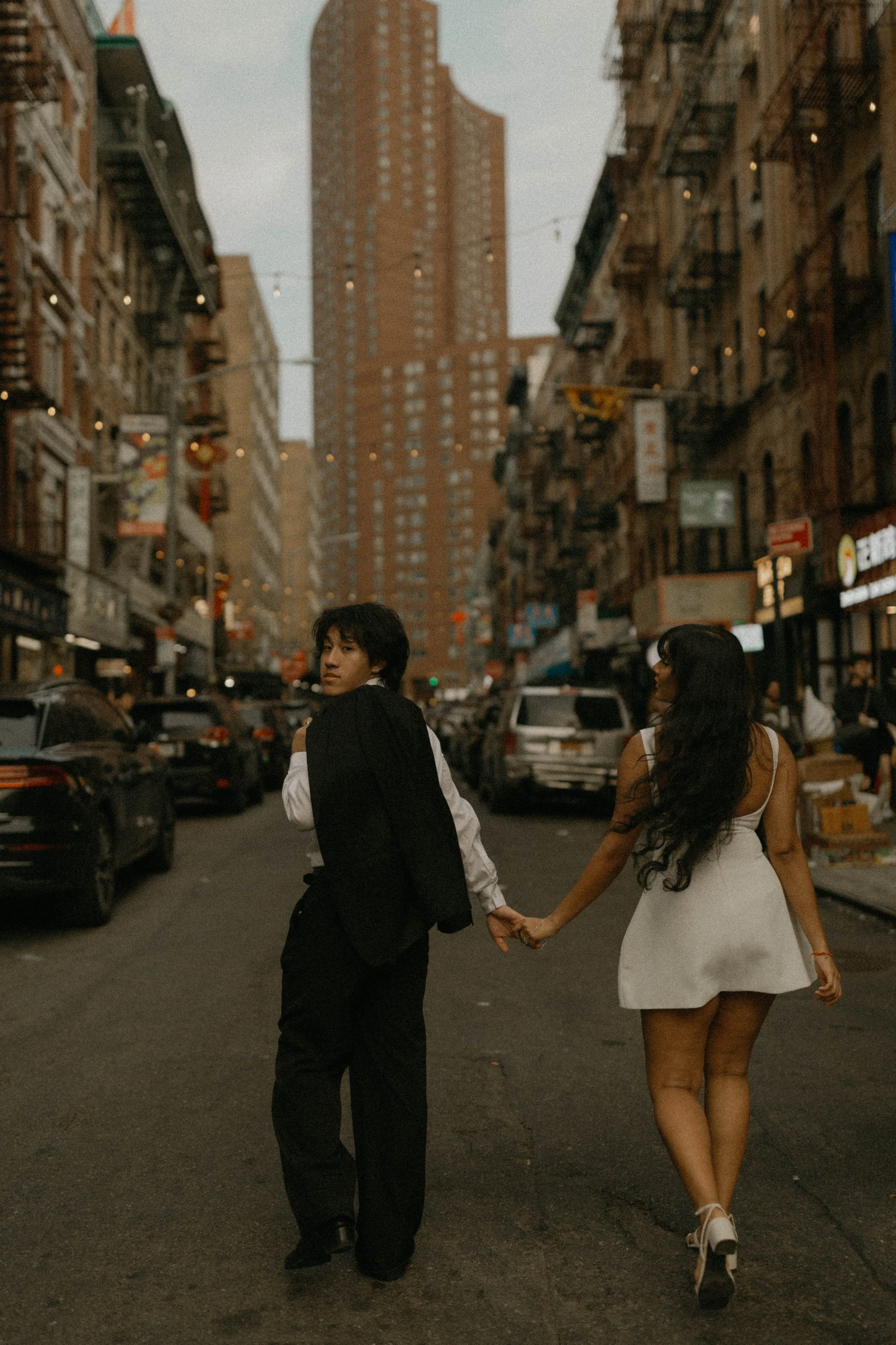 A man and woman holding hands and walking on a city street in the evening, with tall buildings and parked cars around them.