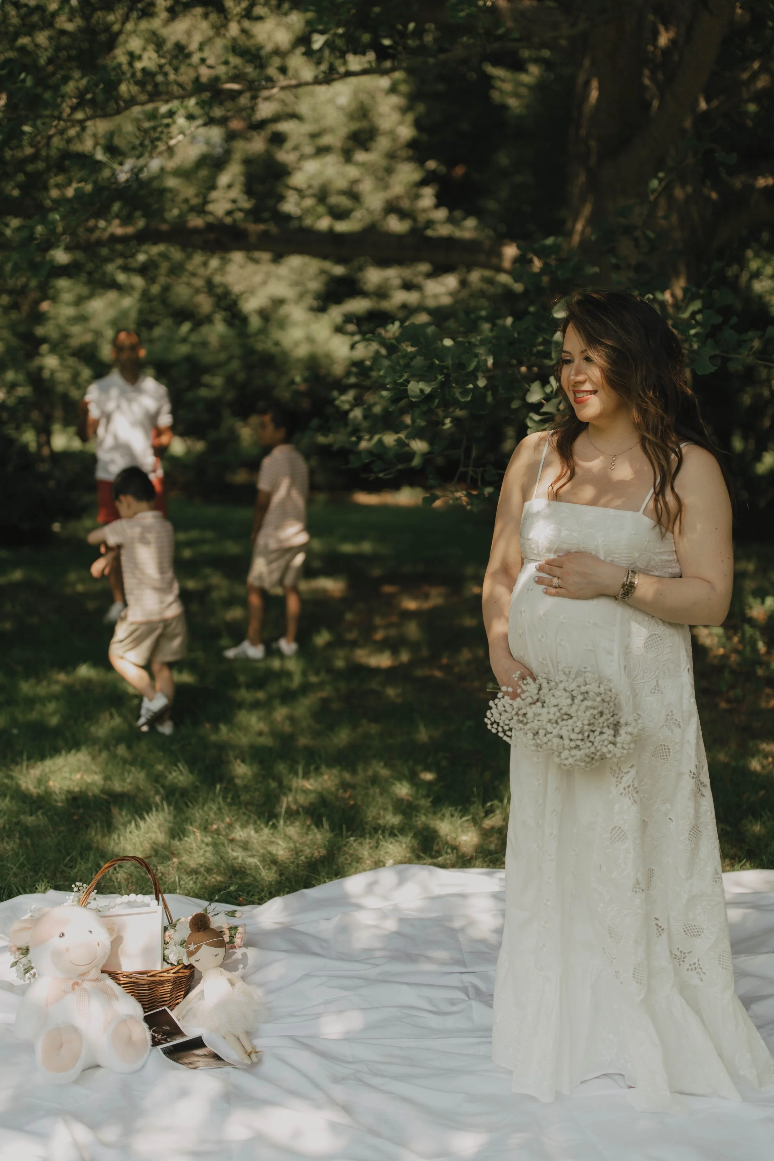 A woman in a white lace dress stands outdoors under trees, holding a bouquet of flowers, smiling. In the background, three children play and a person stands watching. Items like stuffed toys and a basket are arranged on a white cloth on the grass.