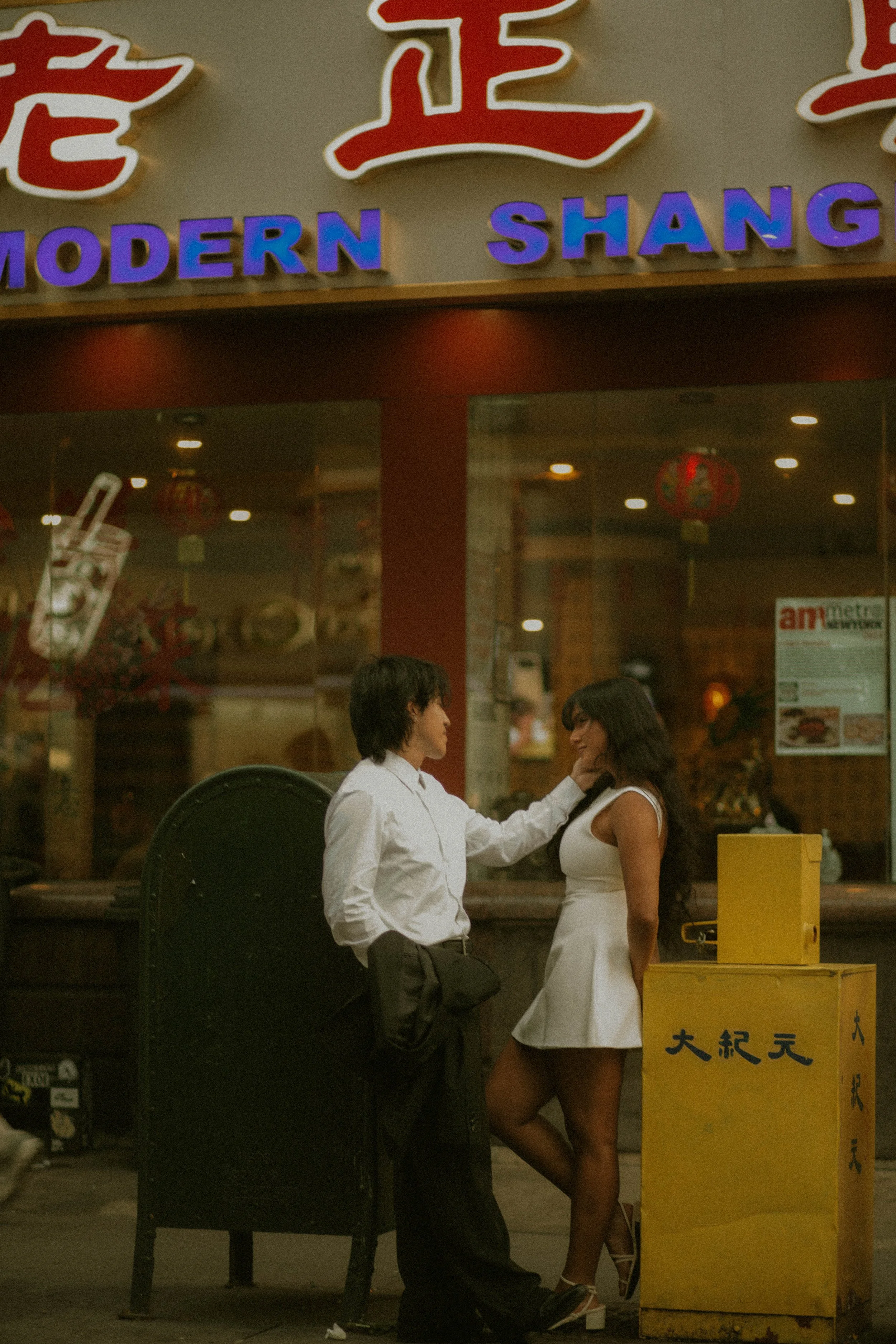 A man and woman standing close outside a restaurant, with the man touching the woman's face; the woman is wearing a white dress, and the man is dressed in a white shirt and dark pants. There are signs and lights in the background.