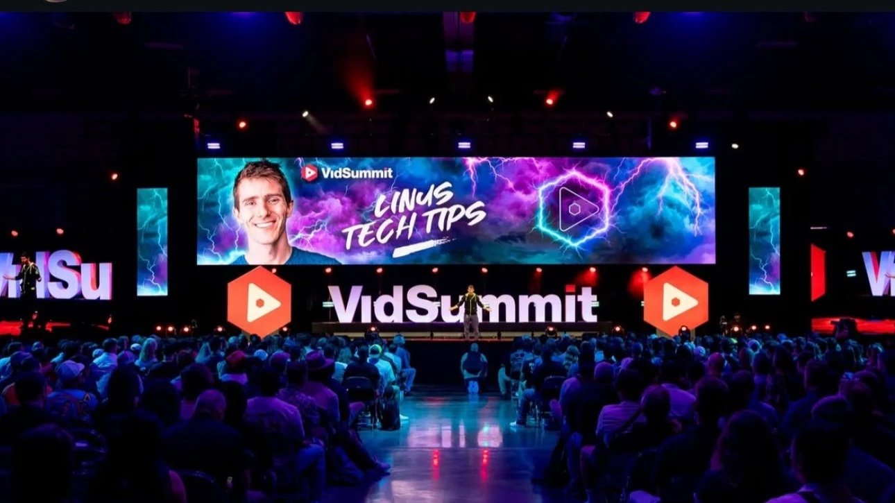 A large conference hall filled with people attending VidSummit. There is a stage with a large screen displaying a presenter with the text 'Linus Tech Tips' and 'VidSummit'. The screen also shows lightning graphics in a purple and blue background. The audience is seated and watching the presentation.