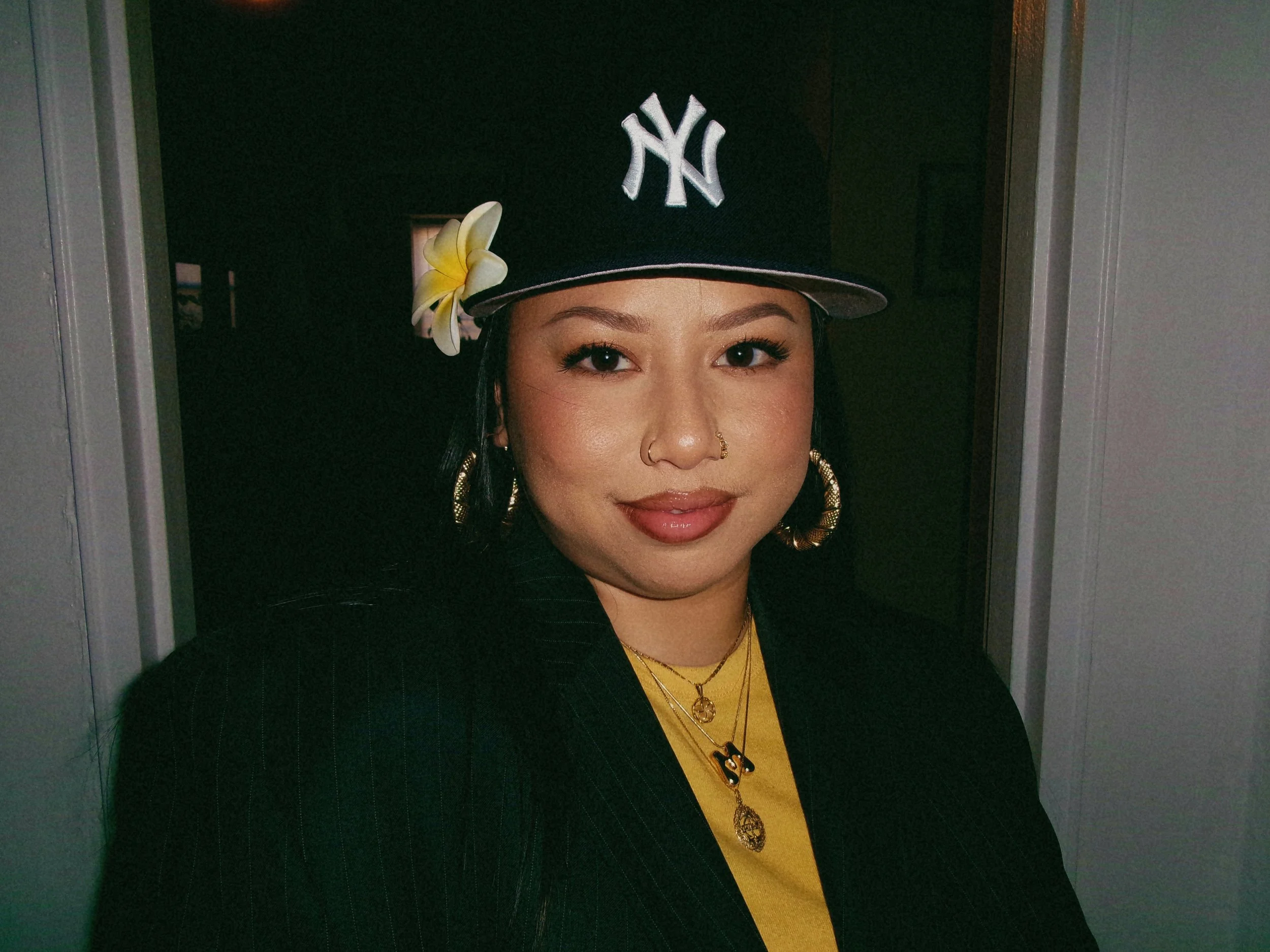 A woman wearing a New York Yankees cap with a yellow flower on the side, gold hoop earrings, layered gold necklaces, and a black blazer, standing indoors with a dark background.
